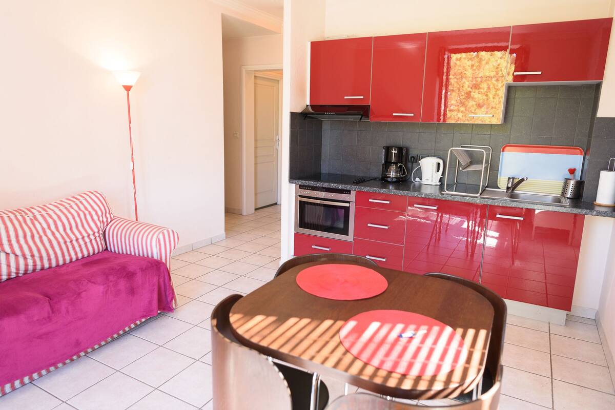 Una cocina compacta con armarios rojos, una mesa de comedor con dos sillas, un sofá con cojines a rayas y una lámpara de pie.
