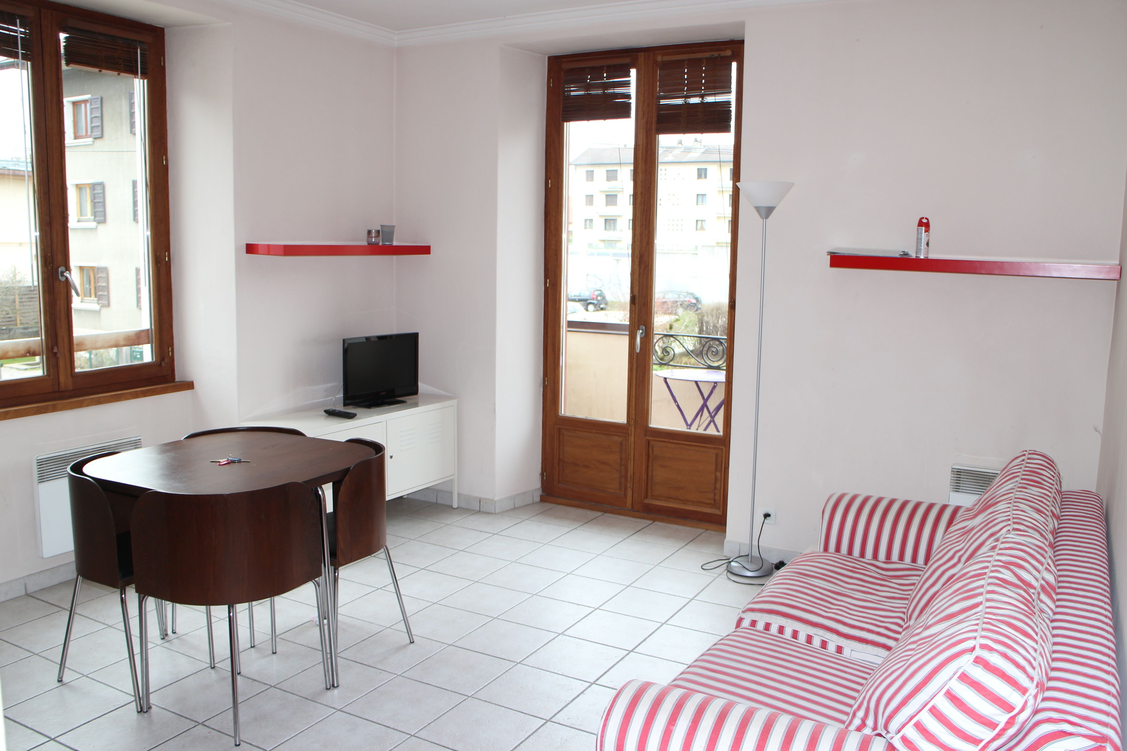 A room with a round table, a sofa, a TV, and a balcony with a view of buildings.