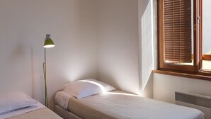 Una cama individual con un almohadón blanco y una lámpara de pie verde en una habitación con ventana y radiador.