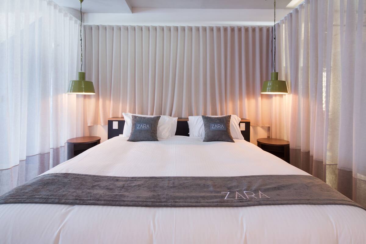 A hotel room with a large bed, two bedside tables, a green pendant lamp, and a Zara-branded pillow.