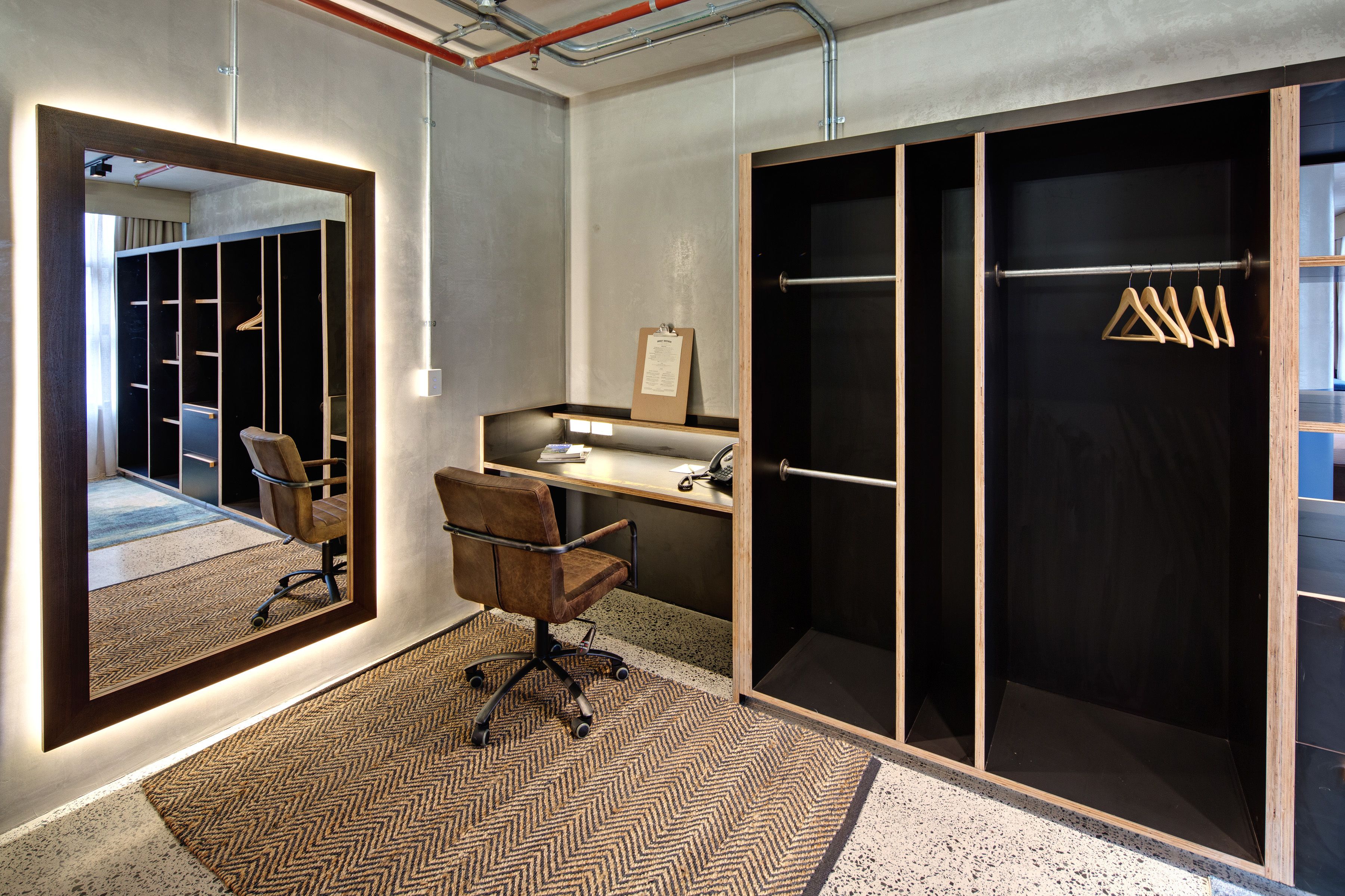 A modern office space with a large mirror, a desk, a chair, and a wardrobe with hanging clothes.