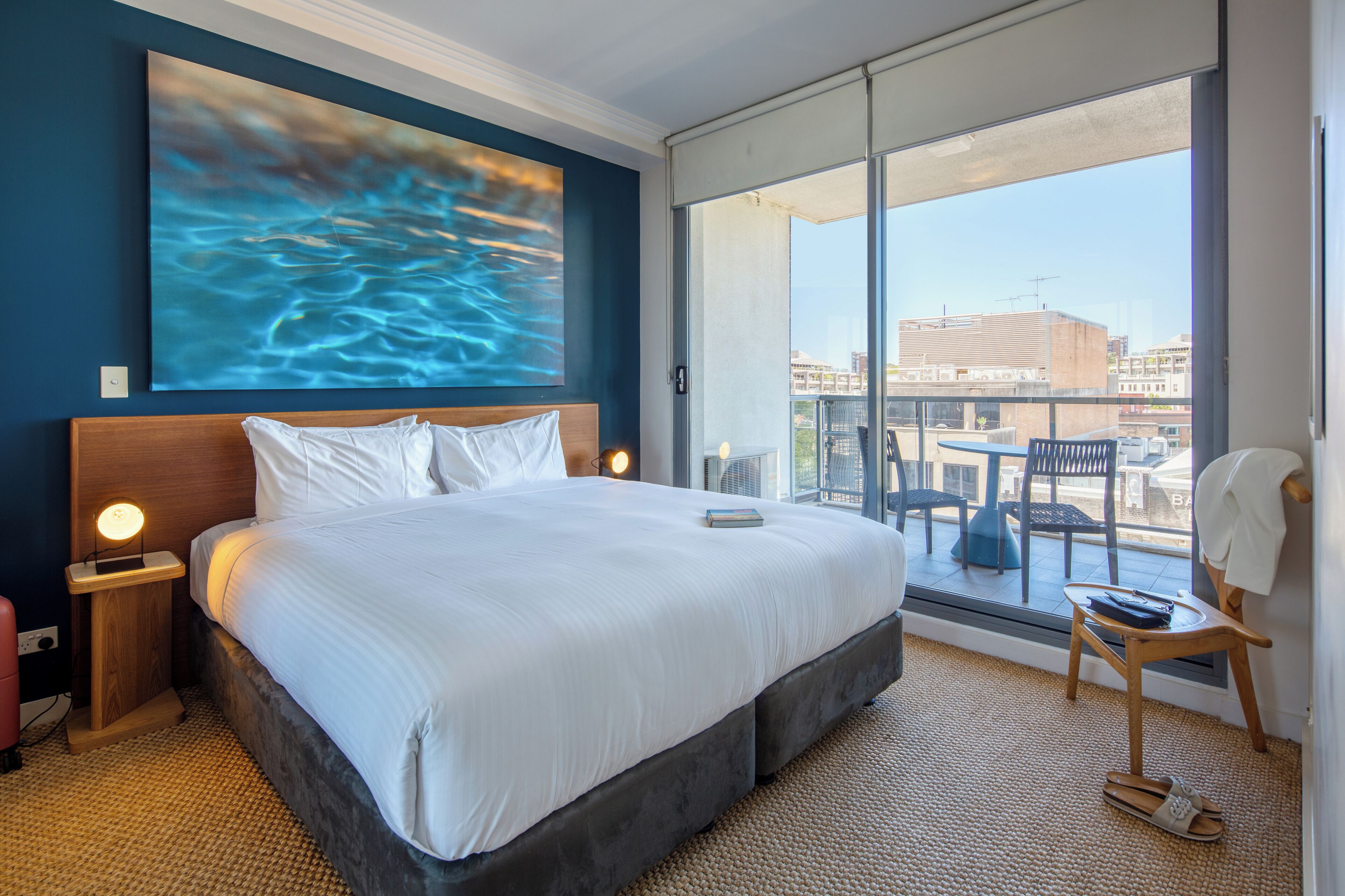 Quarto de hotel com uma cama grande, vista da cidade através de uma porta deslizante e uma varanda com mesa e cadeiras.