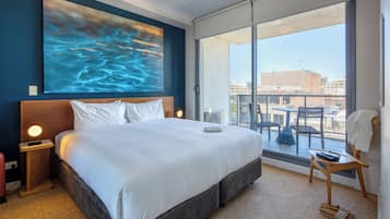 Quarto de hotel com uma cama grande, vista da cidade através de uma porta deslizante e uma varanda com mesa e cadeiras.