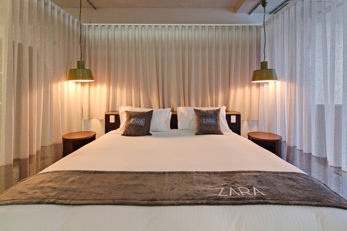 A hotel room with a large bed, two bedside tables, a headboard, and two hanging lamps.