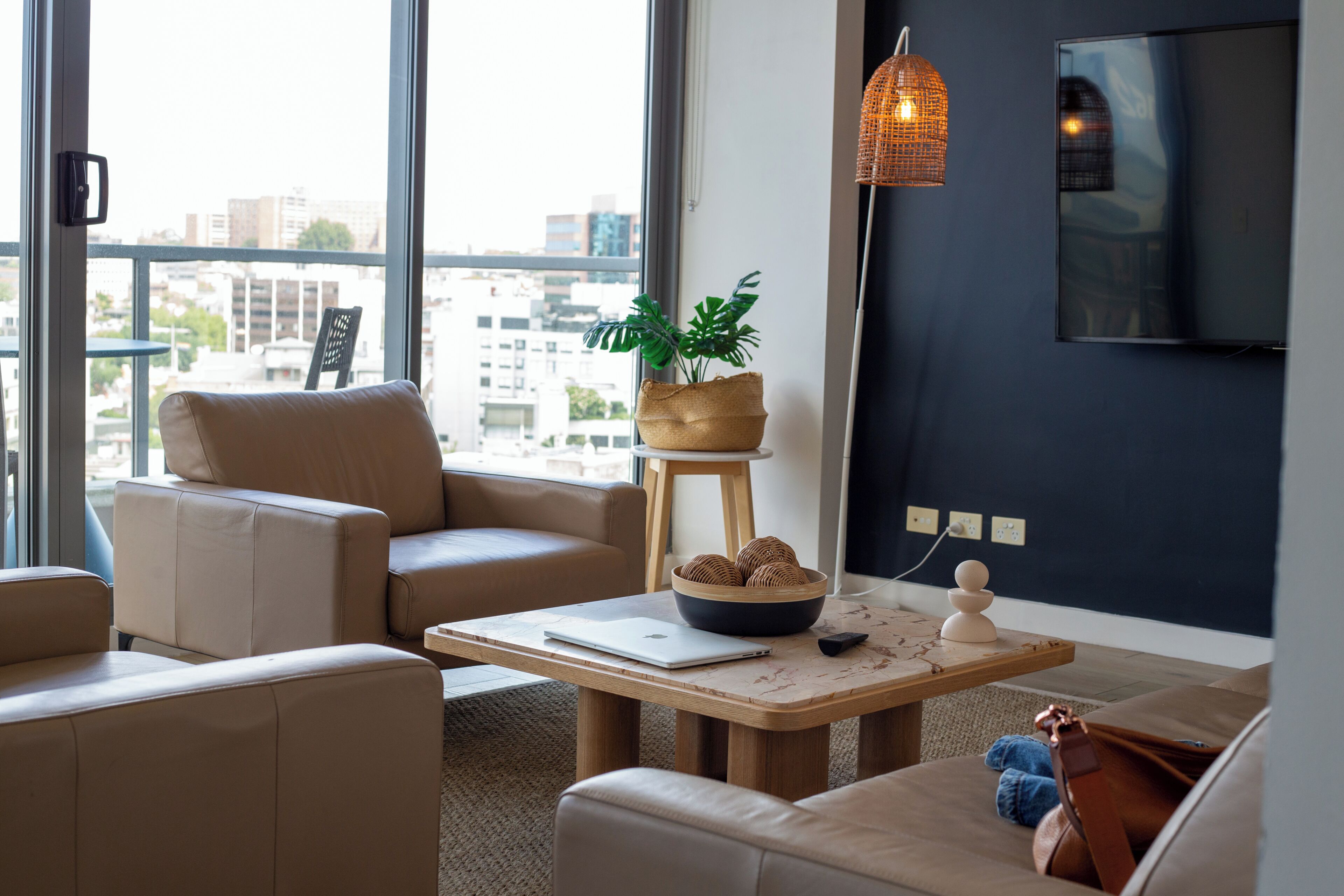 Una sala de estar moderna con un amplio ventanal, un ordenador portátil sobre una mesa, una planta y un televisor montado en la pared.