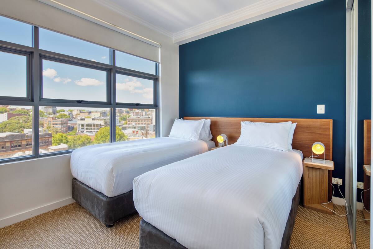 A hotel room with two beds, a view of the city, and a dark blue accent wall.