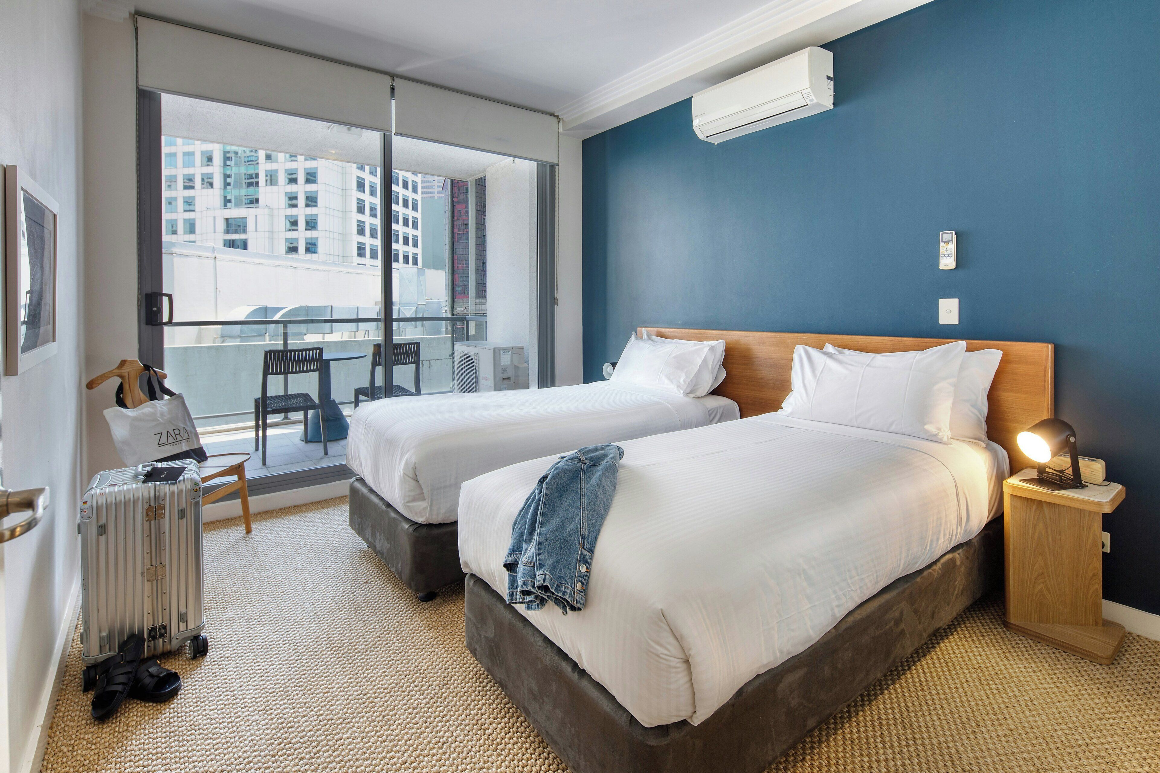A hotel room with two beds, a balcony with a view of buildings, and a luggage trolley.