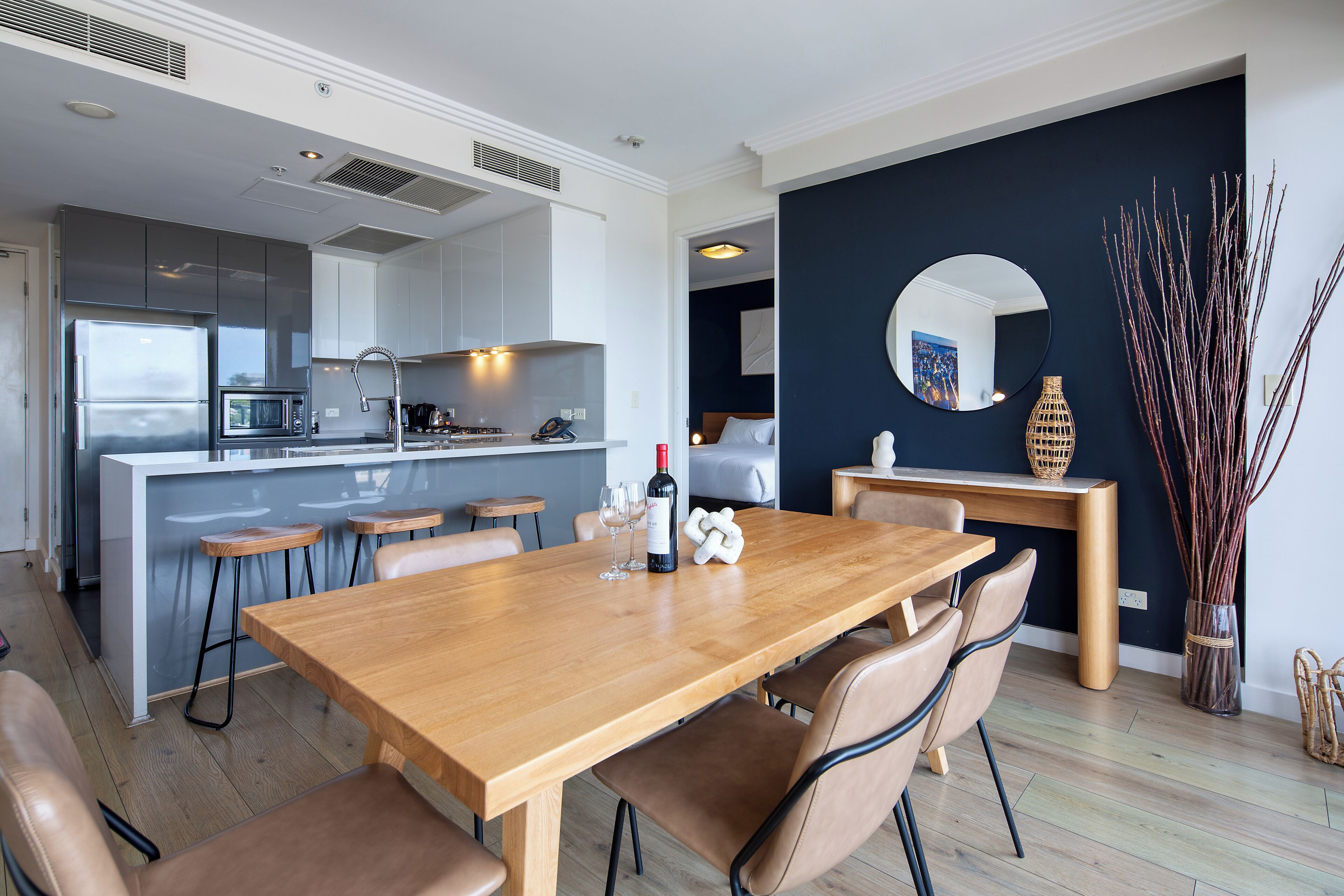 Une cuisine moderne dotée d’un îlot central, d’un coin repas avec une table et des chaises en bois, et d’un mur d’accent de couleur sombre agrémenté d’un miroir rond.