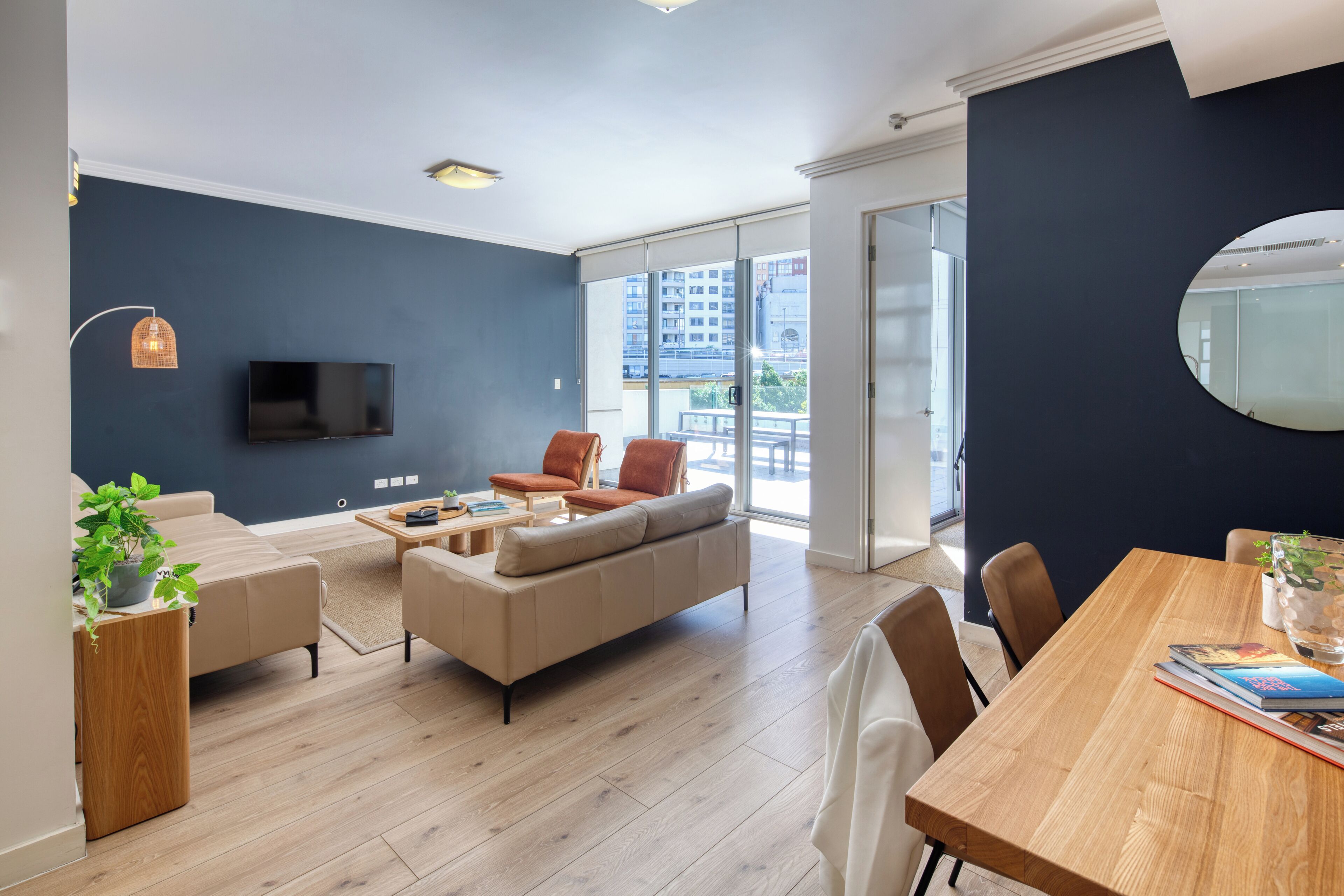 Una sala de estar moderna con un sofá, una mesa de centro y una mesa de comedor, un televisor montado en la pared, y un ventanal con vistas a edificios.