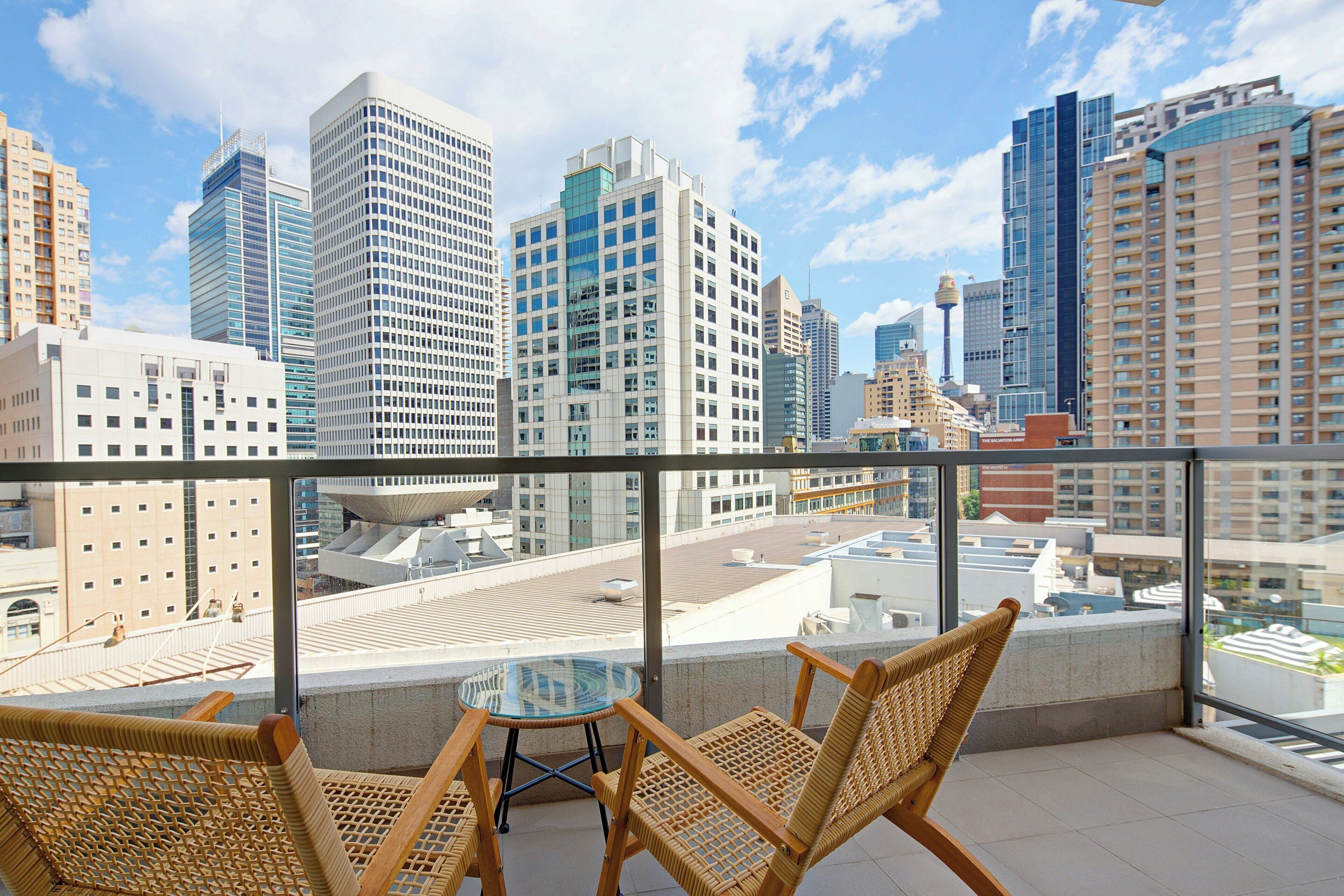 A balcony with wicker chairs and a glass table overlooking a cityscape with modern buildings.