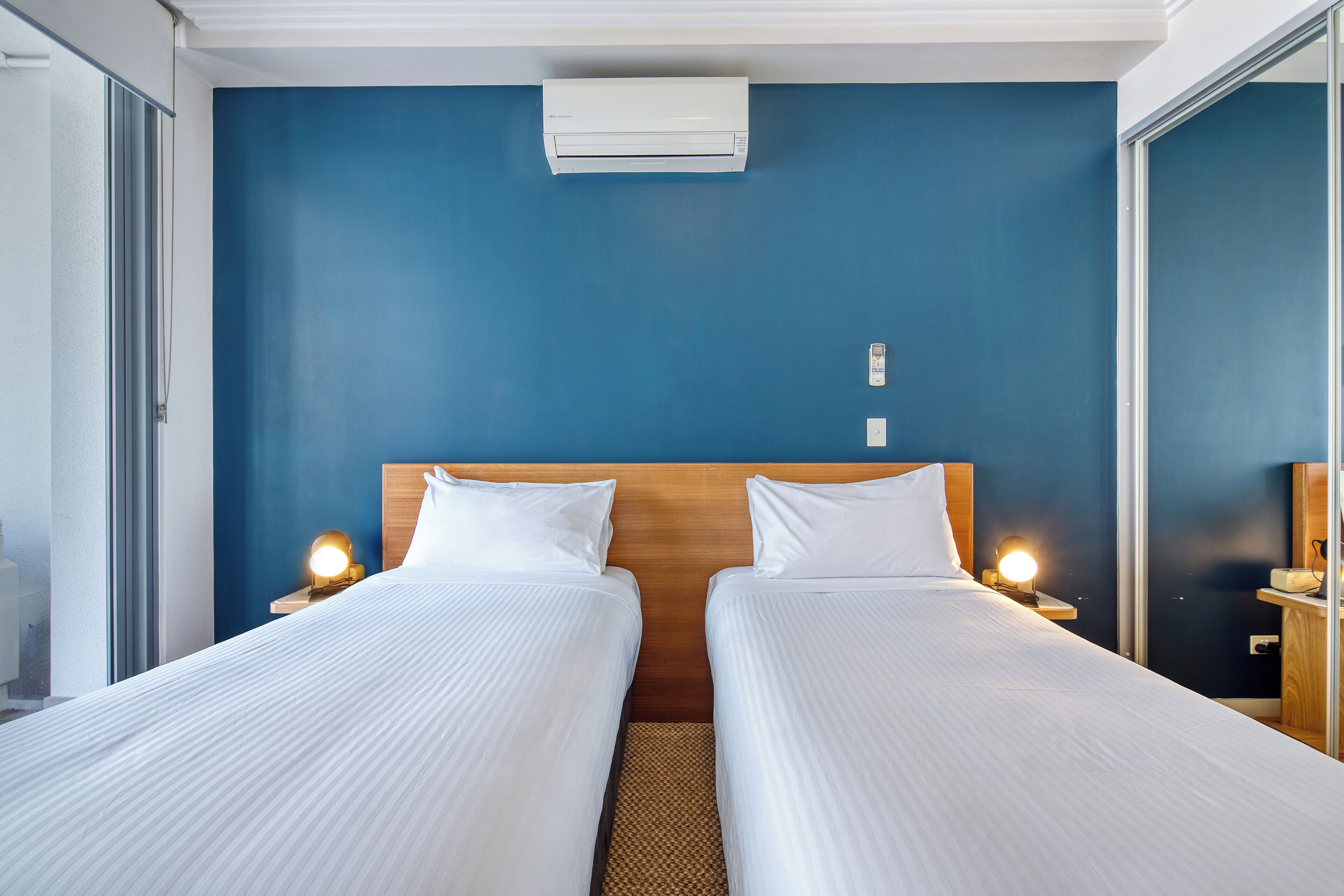 A hotel room with two beds, a wooden headboard, bedside lamps, an air conditioner, and a window.