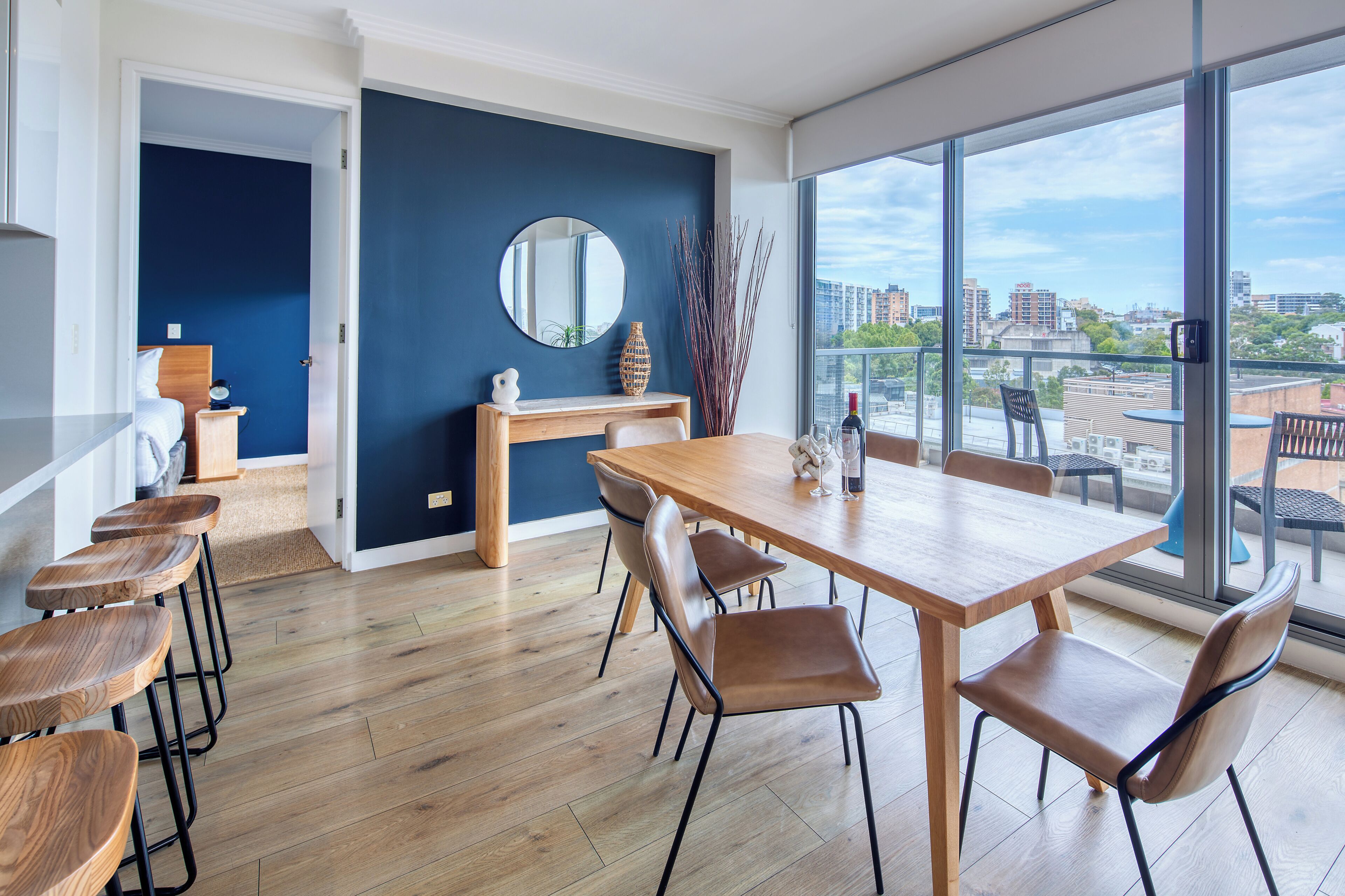 A modern dining area with a wooden table and chairs, a round mirror, and a view of the city through large windows.