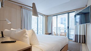 A modern hotel room with a large bed, a desk with a lamp, a TV mounted on the wall, and a view of the city through large windows. - Zara Tower Hotel (Sydney)