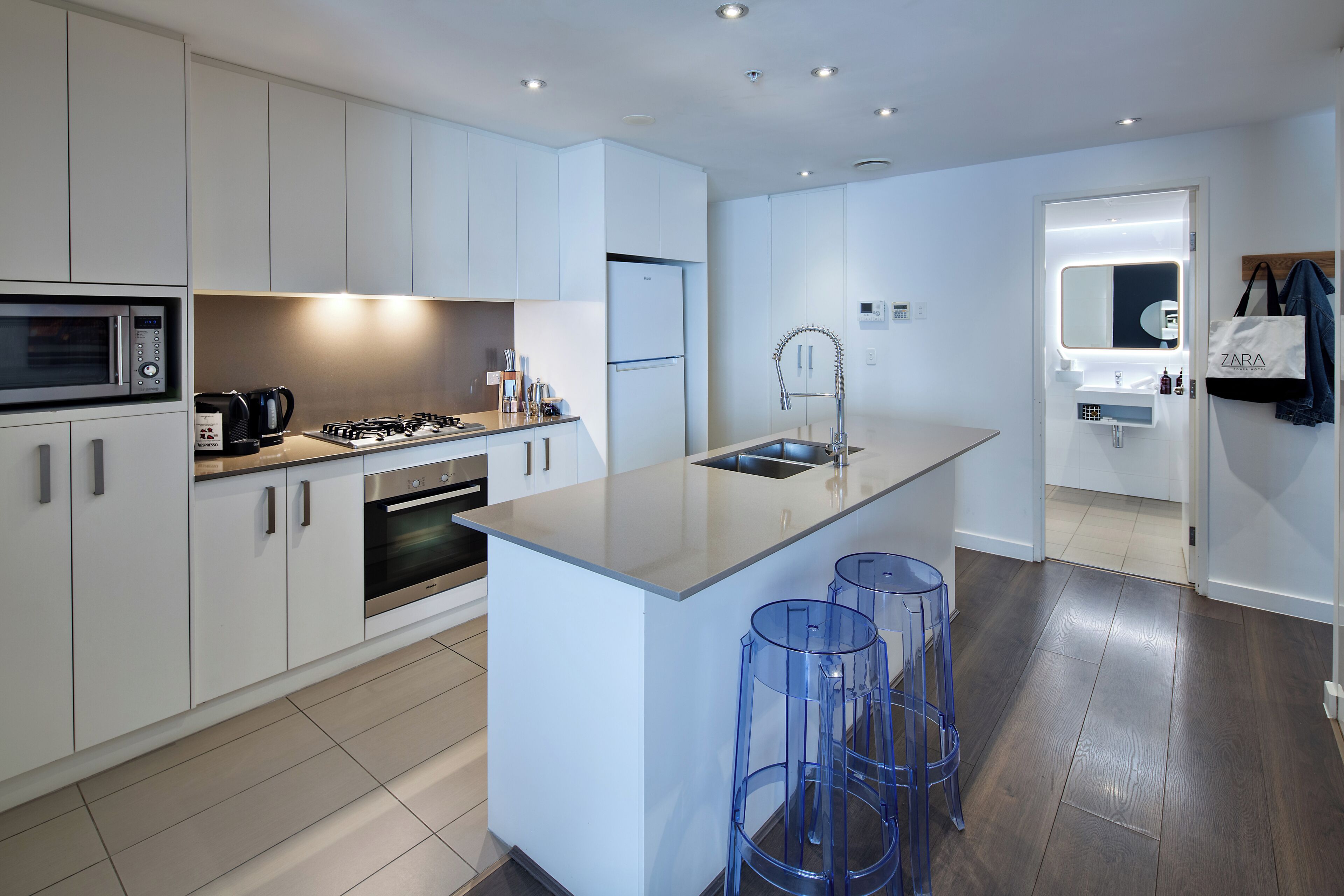 Una cocina moderna con armarios blancos, una isla central, taburetes de bar y un baño a la vista con espejo y perchero.