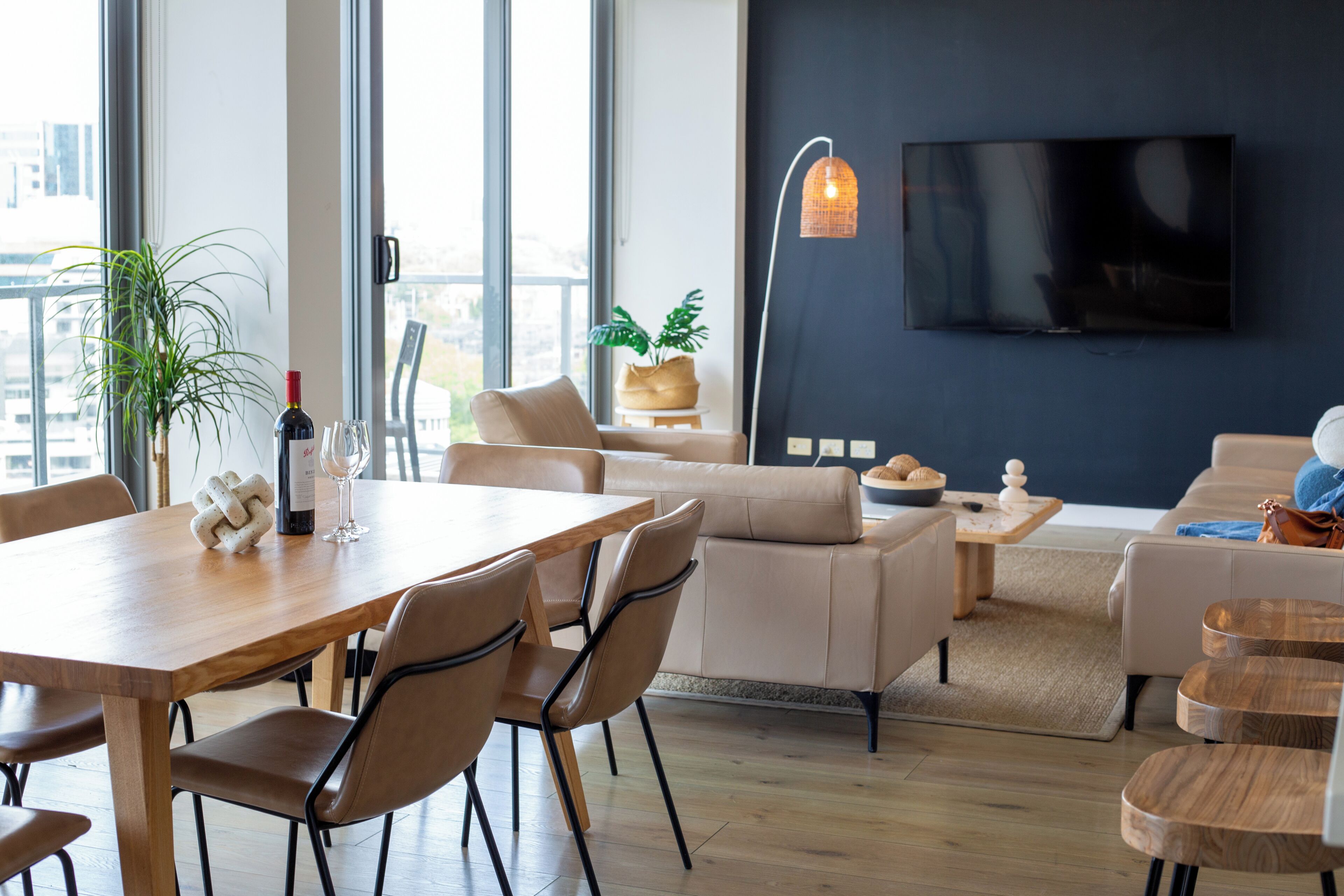 A modern living room with a dining table, chairs, sofas, a TV mounted on the wall, and a large window.