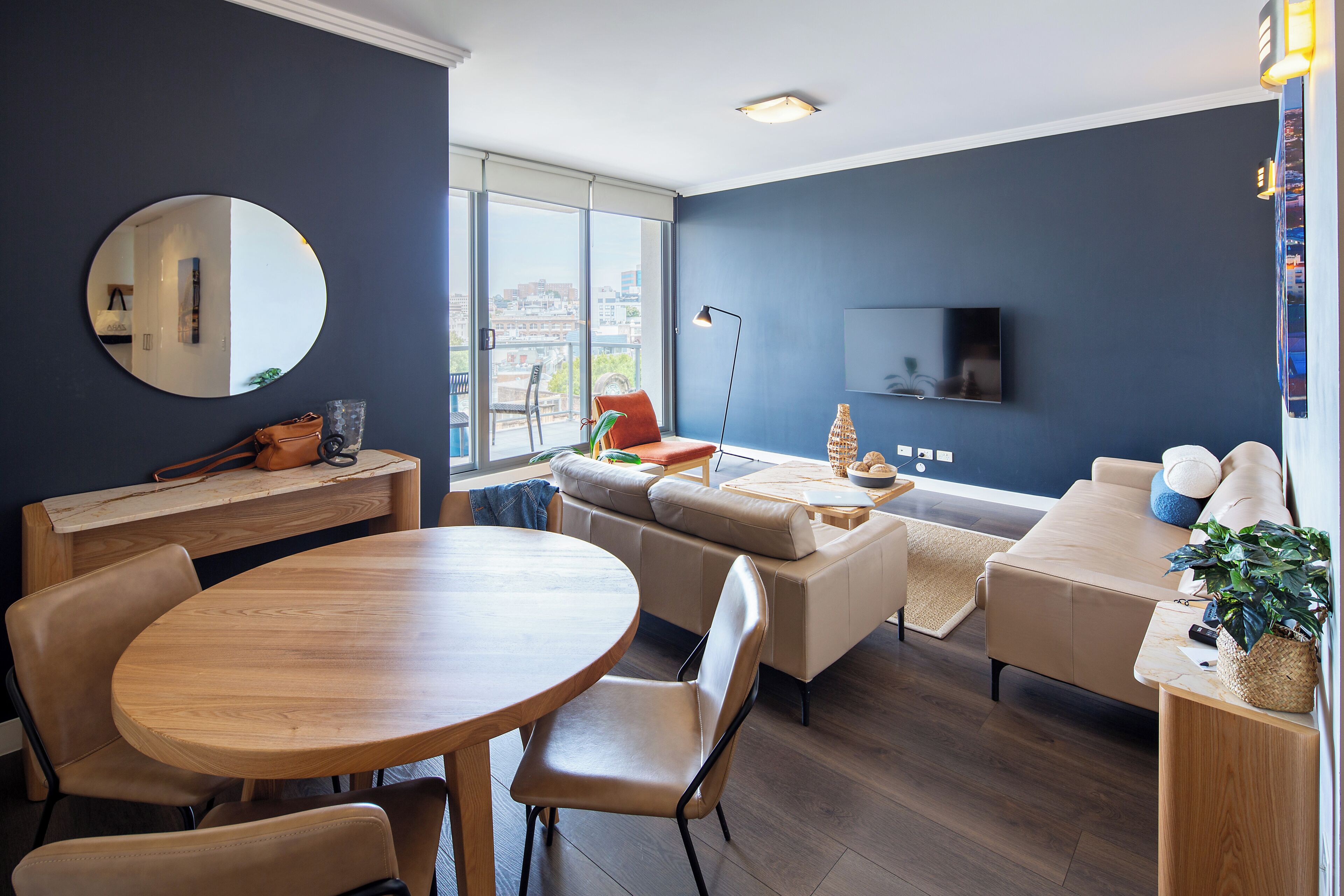 Un salon moderne avec une table ronde en bois, des canapés beiges, un grand miroir et un téléviseur à écran plat fixé au mur.