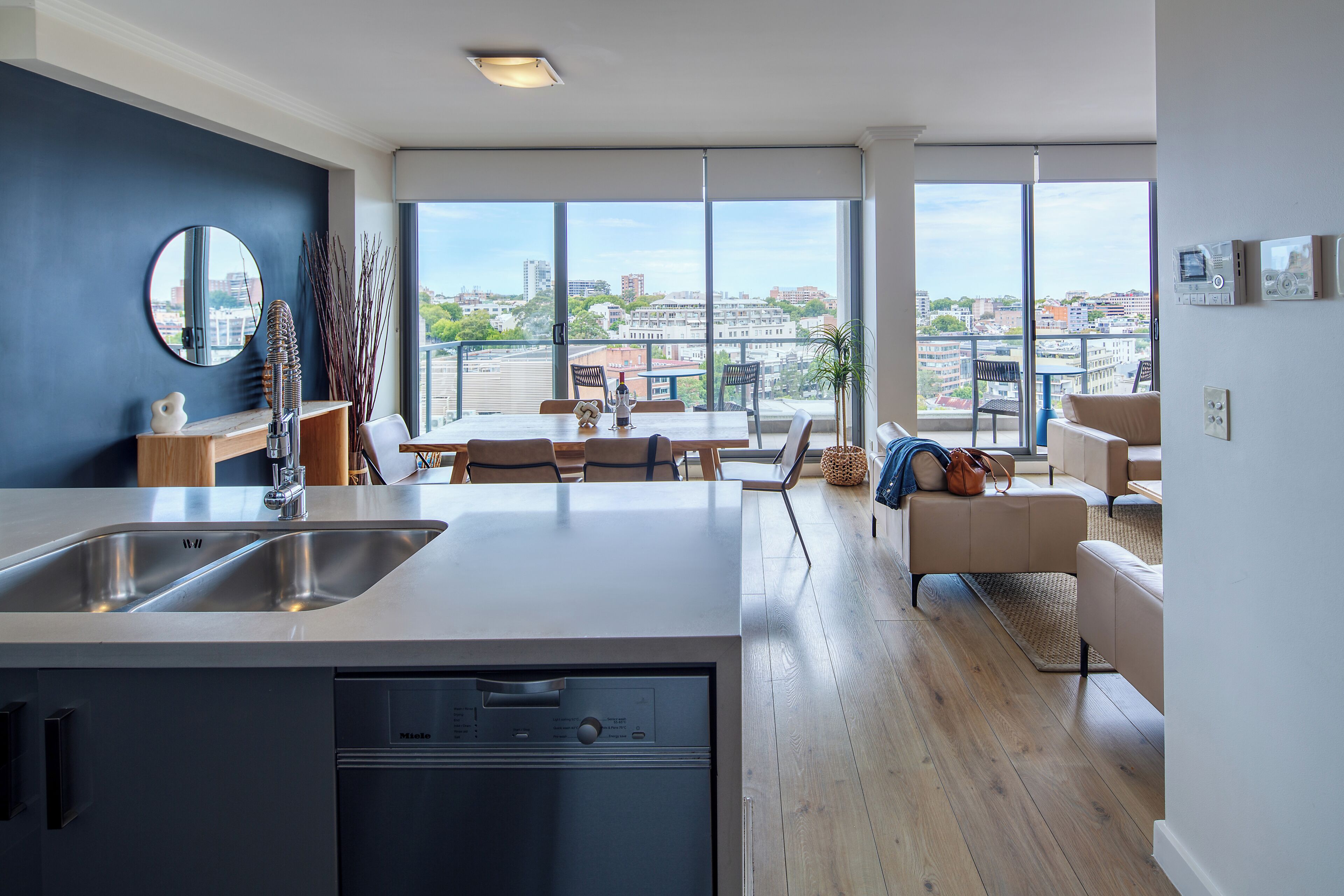 Una cocina moderna con lavavajillas, un comedor con mesa y sillas, y un amplio ventanal con vista a la ciudad.