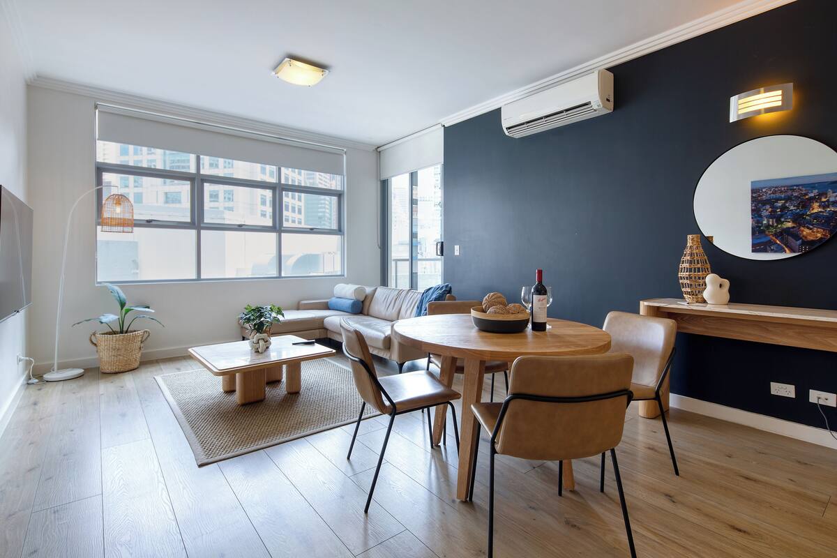 A modern living room with a sofa, dining table, chairs, a large window, and a wall-mounted air conditioner.