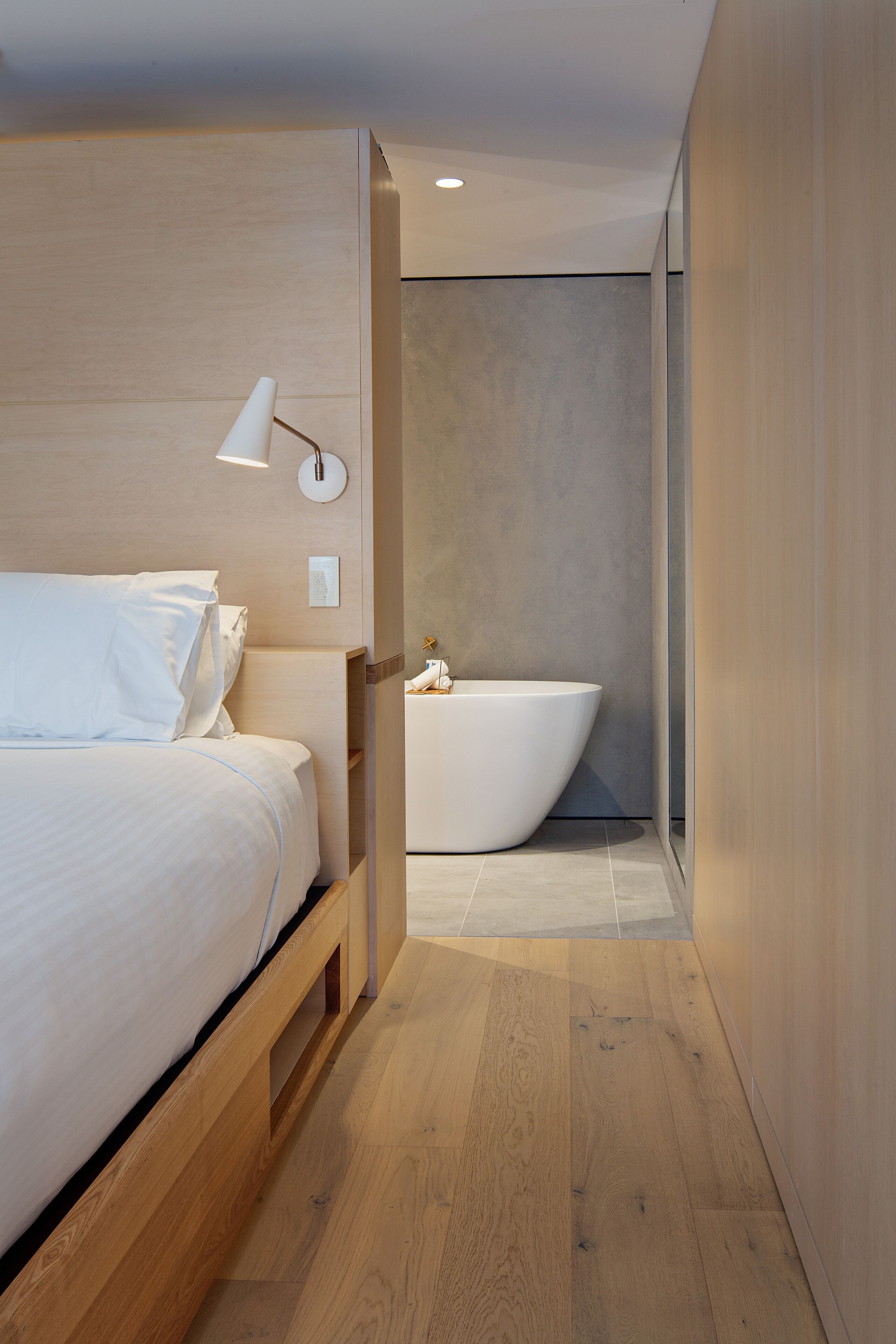 A modern hotel room with a wooden bed, a white bedside table, a wall-mounted lamp, and a large bathtub.