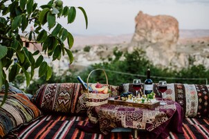 A picnic setup with a basket of fruits, cheese, and wine on a patterned blanket, overlooking a mountainous landscape.