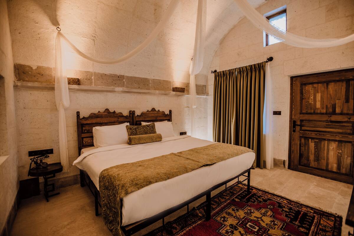 A bedroom with a large bed, a wooden headboard, a nightstand, a patterned rug, and a wooden door.