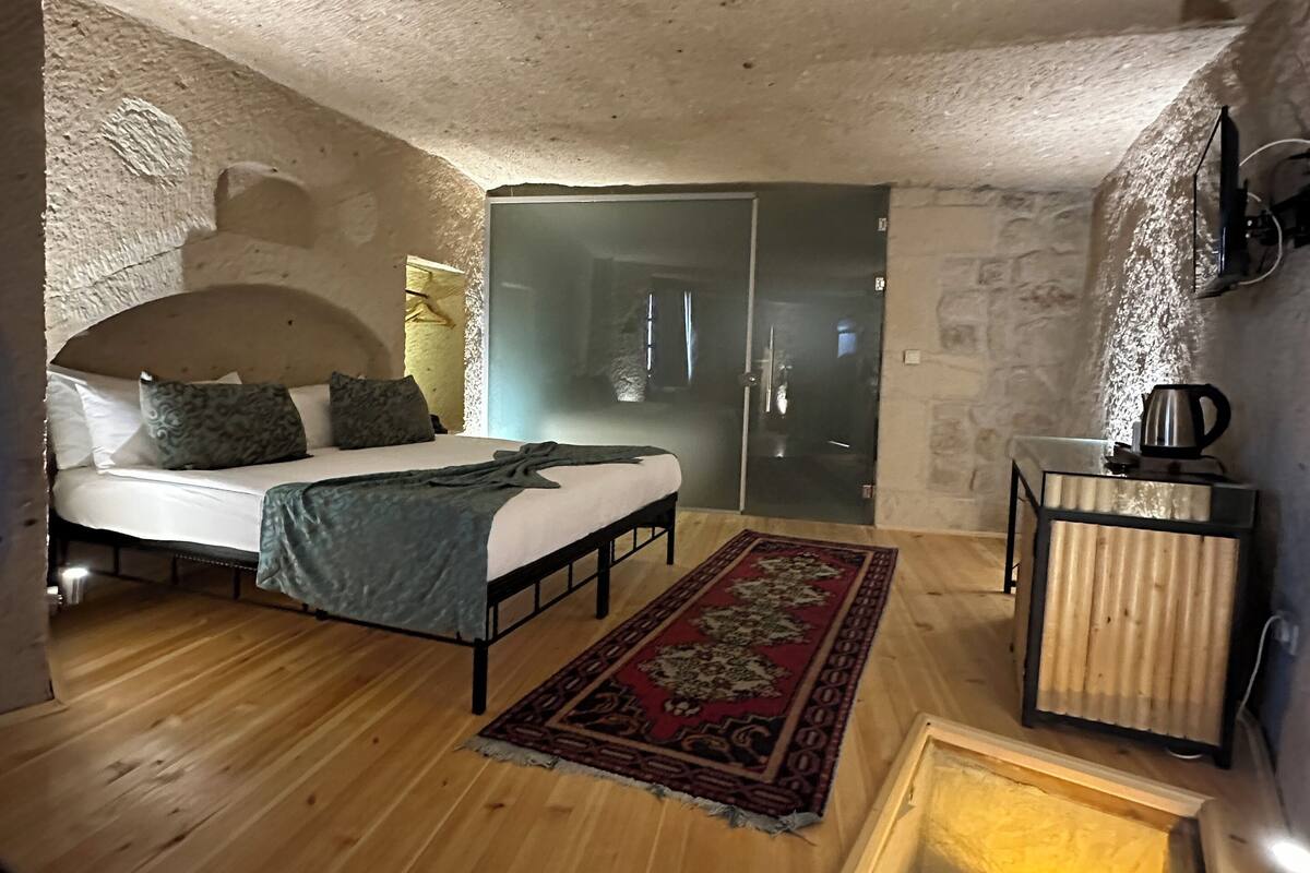 A bedroom with a bed, a wooden floor, a red patterned rug, a small wooden cabinet, and a wall-mounted TV.