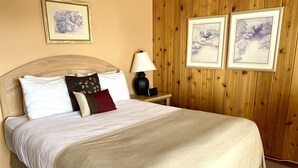 Cama bem arrumada com colcha bege e almofadas, cabeceira de madeira, mesa de cabeceira com abajur e duas imagens emolduradas na parede.