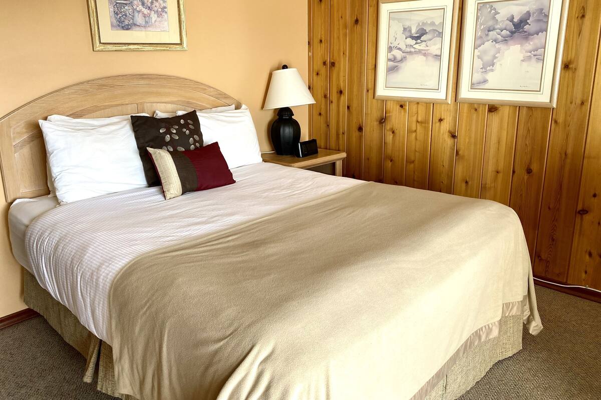 Een netjes opgemaakt bed met een beige sprei en kussens, een houten hoofdbord, een nachtkastje met een lamp, en twee ingelijste foto’s aan de muur.