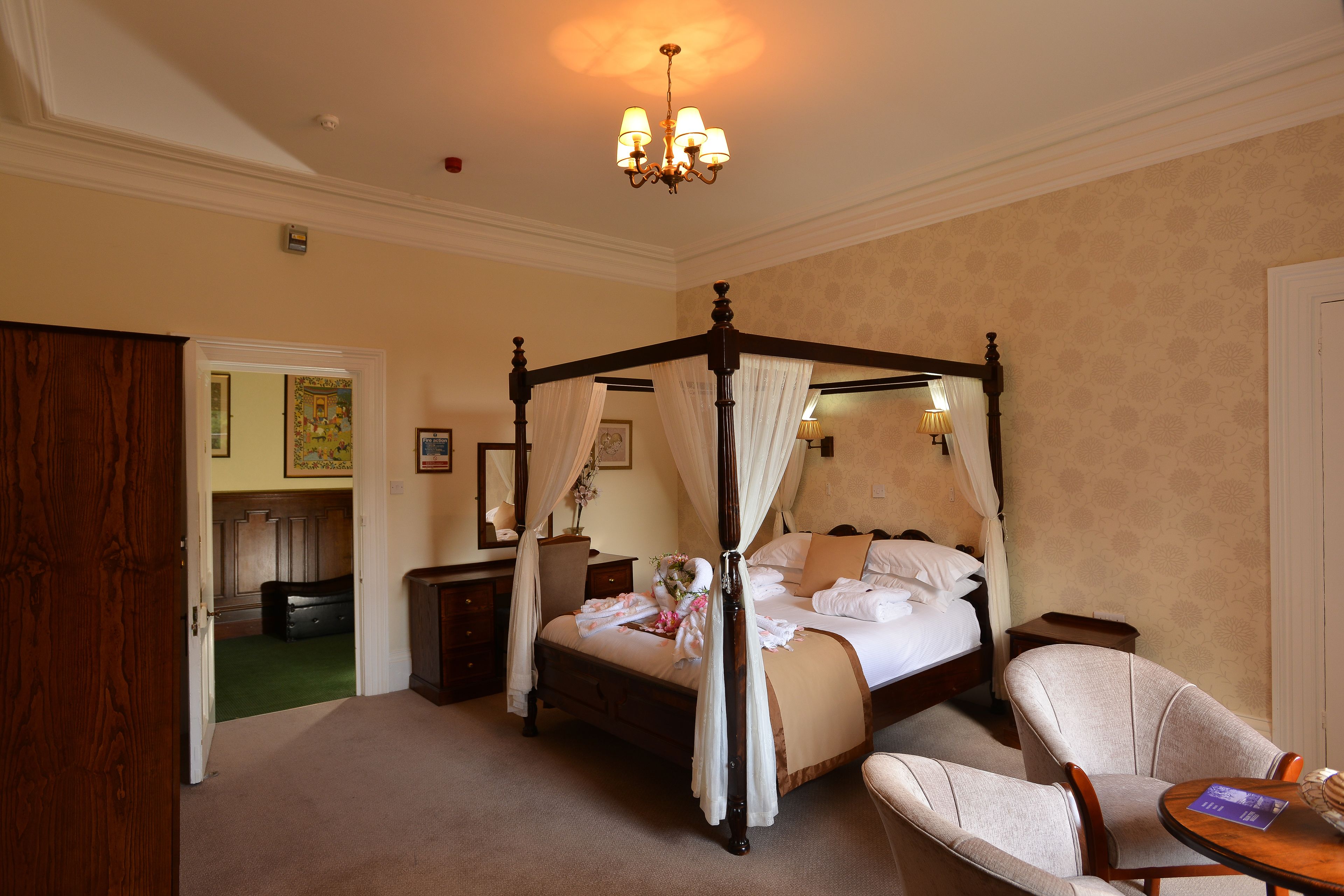 A four-poster bed with a canopy, a sitting chair, a bedside table, and a small round table with a lamp.
