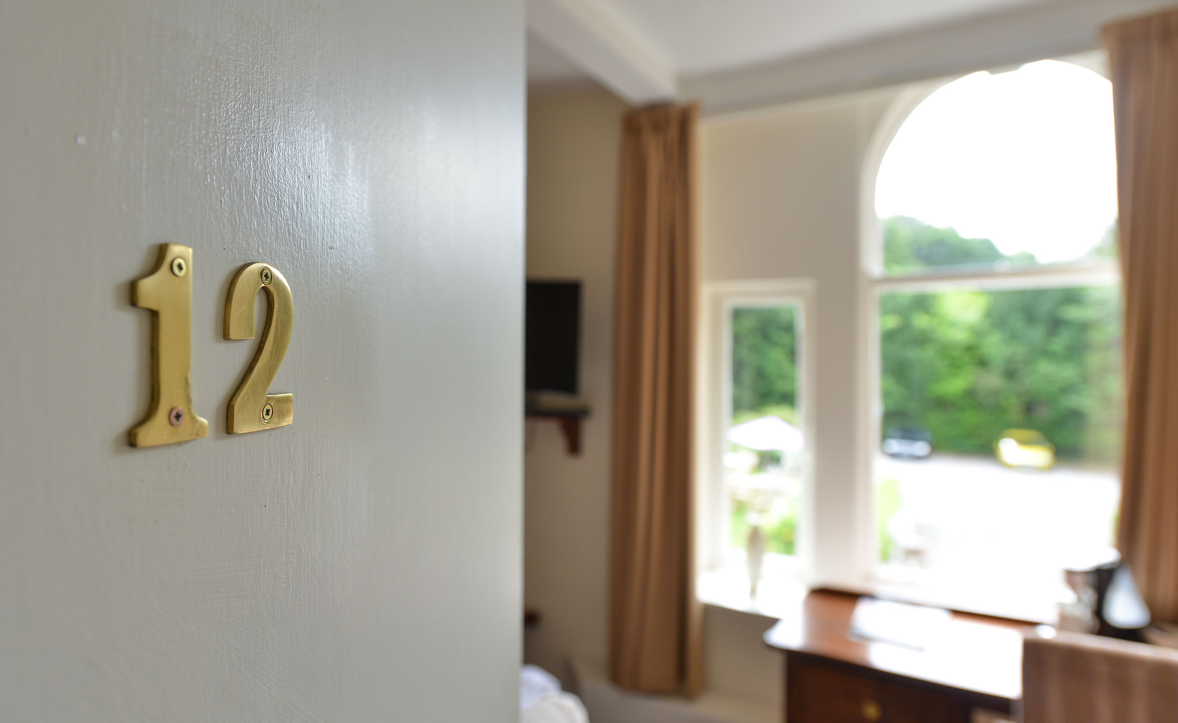 A room with a number '12' on the wall, a window with curtains, and a view of greenery outside.