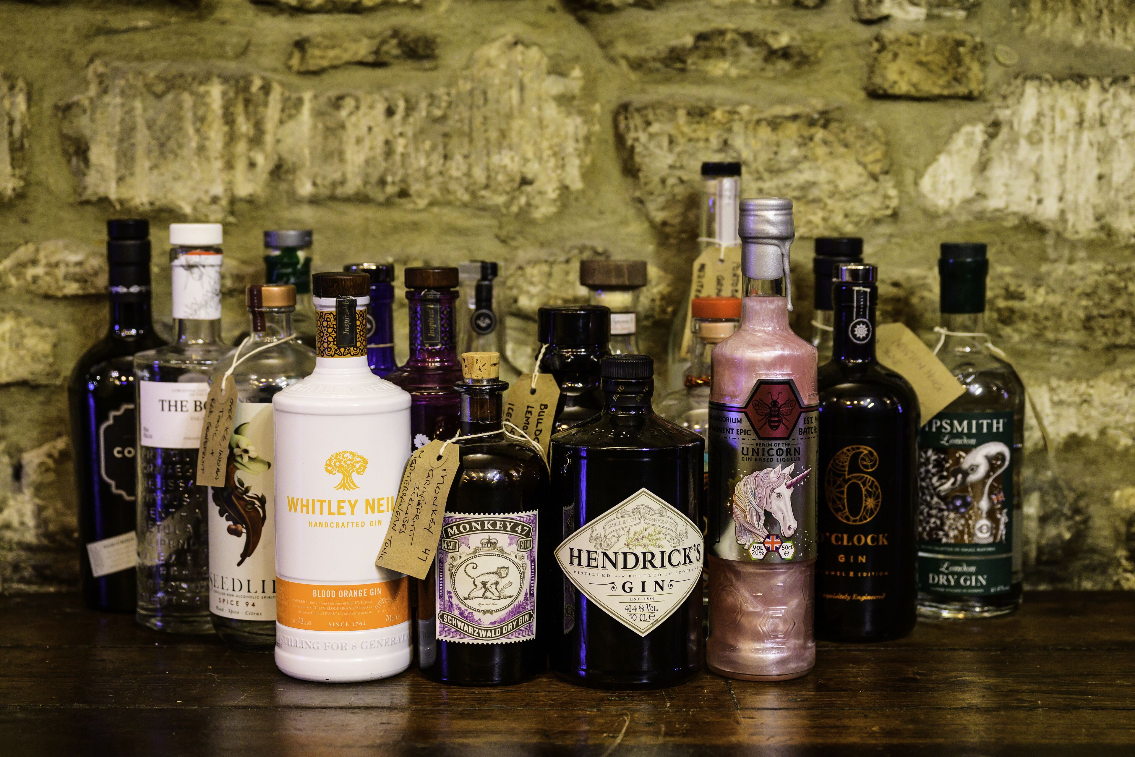 A variety of gin bottles displayed on a wooden surface against a stone wall.