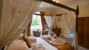 A bedroom with a four-poster bed, a sitting area with chairs, and a view of the outdoors. - Steeton Hall Hotel (Keighley)