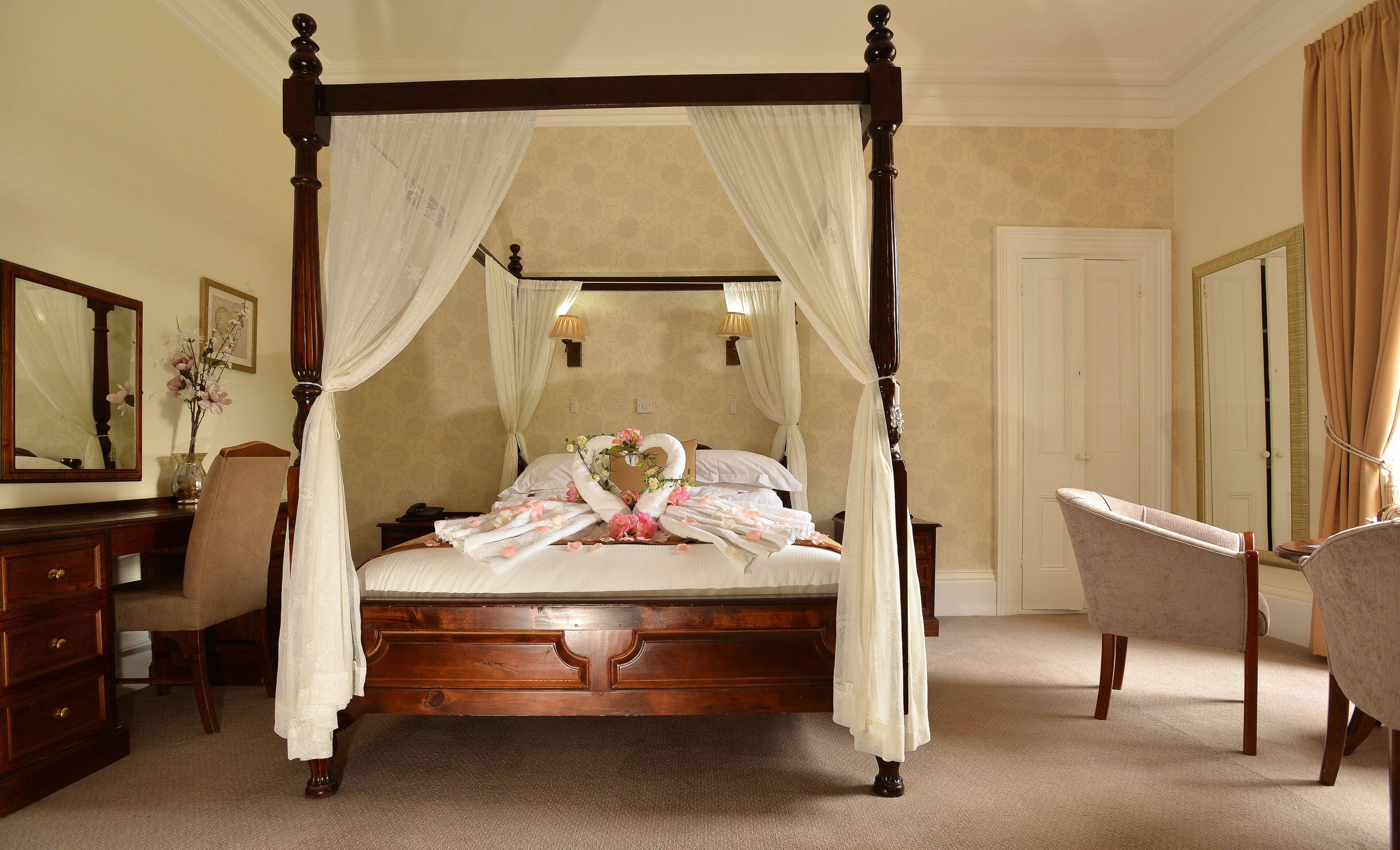 A four-poster canopy bed with a floral arrangement, a mirror, and a chair in a room with beige walls and carpet.