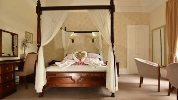 A four-poster canopy bed with a floral arrangement, a mirror, and a chair in a room with beige walls and carpet.