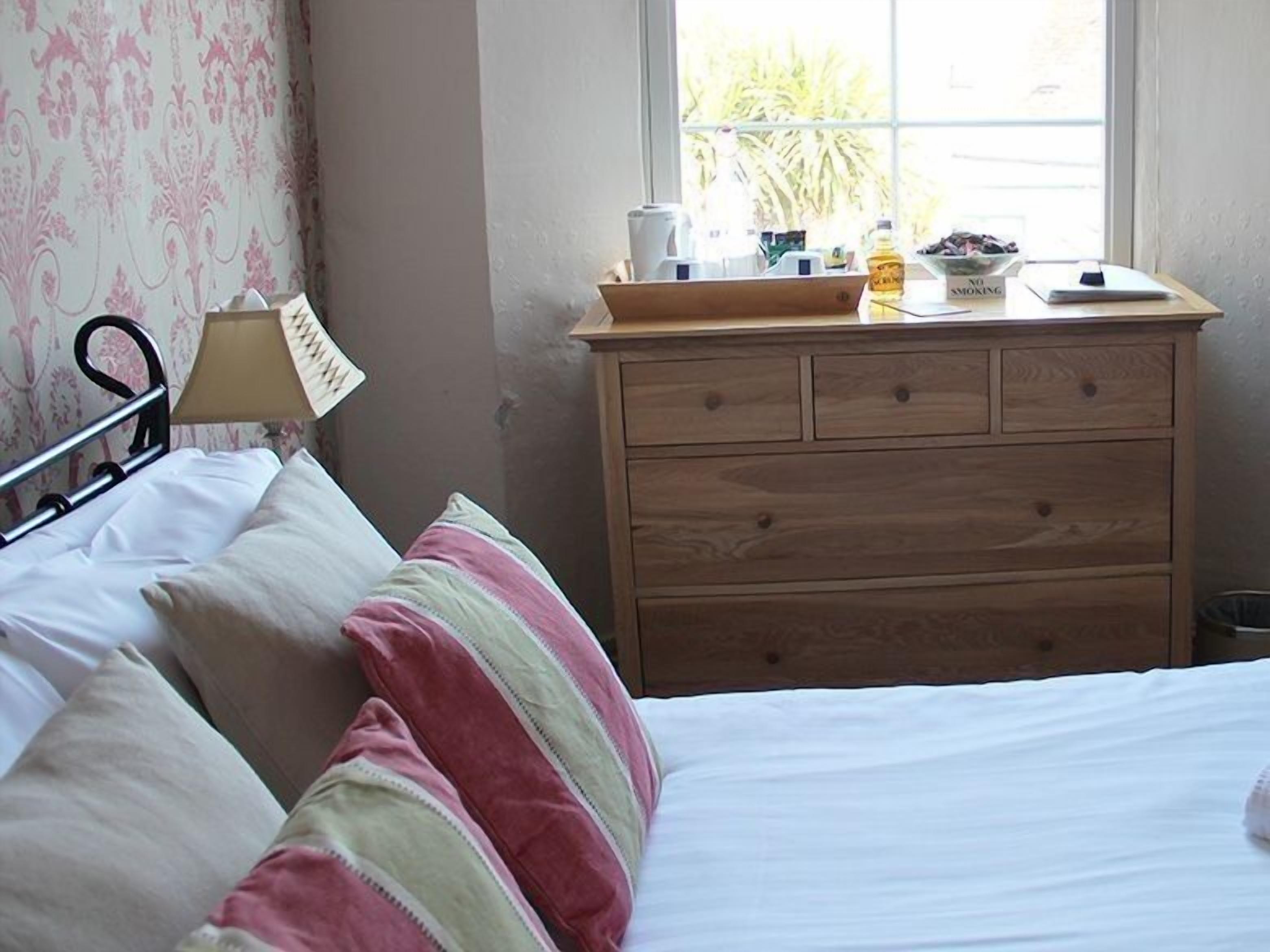 A bedroom with a bed, a wooden dresser, a window with a view of greenery, and a lamp on the dresser.