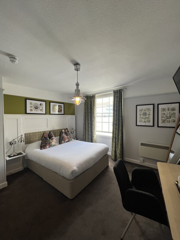Habitación de hotel con cama, mesita de noche, lámpara, silla, escritorio y ventana con cortinas.