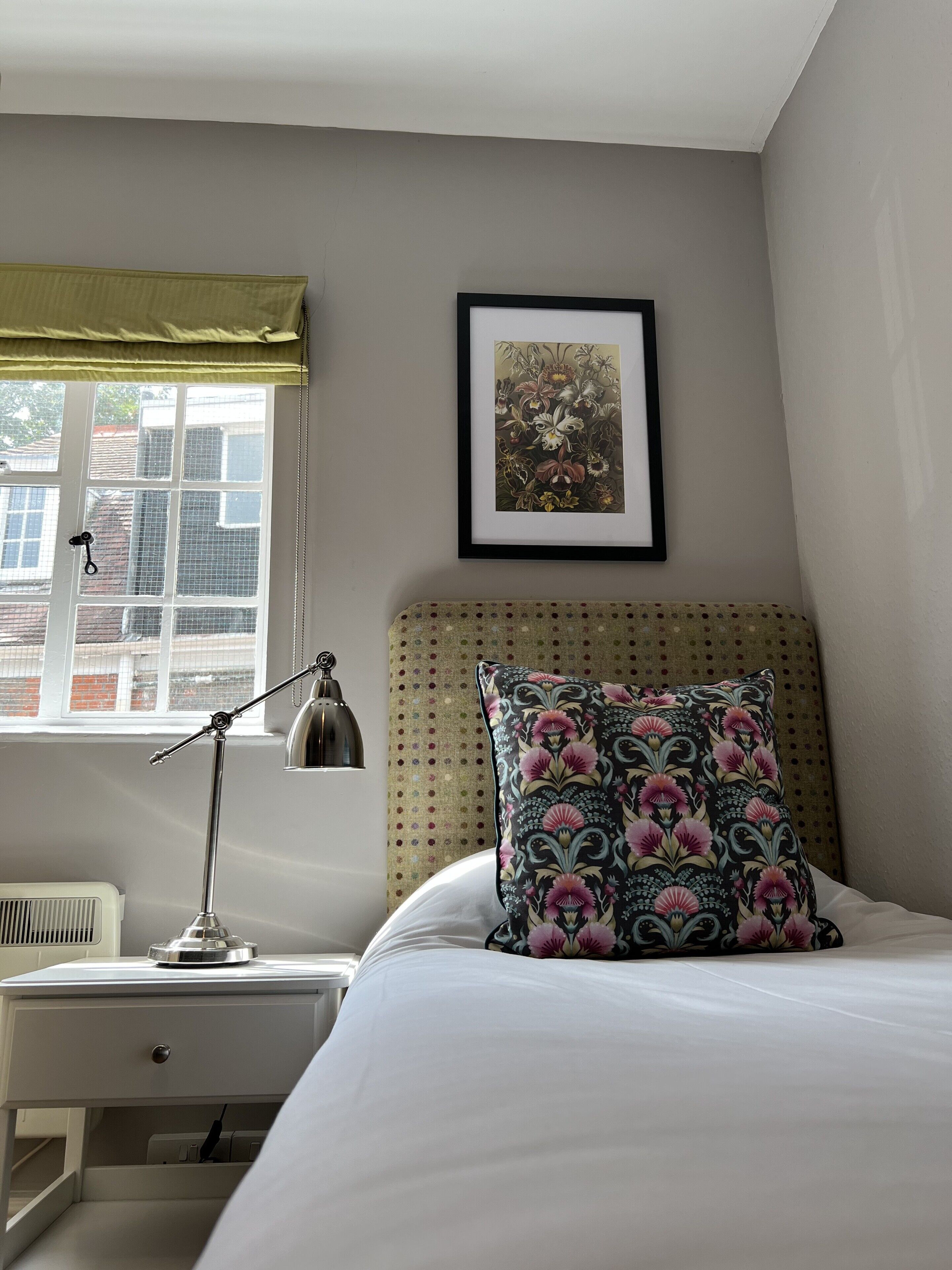 Un dormitorio con cama, mesita de noche, lámpara, ventana con vistas a edificios y un cuadro en la pared.