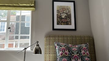 Ein Schlafzimmer mit Bett, Nachttisch, Lampe, Fenster mit Blick auf Gebäude und einem gerahmten Bild an der Wand.