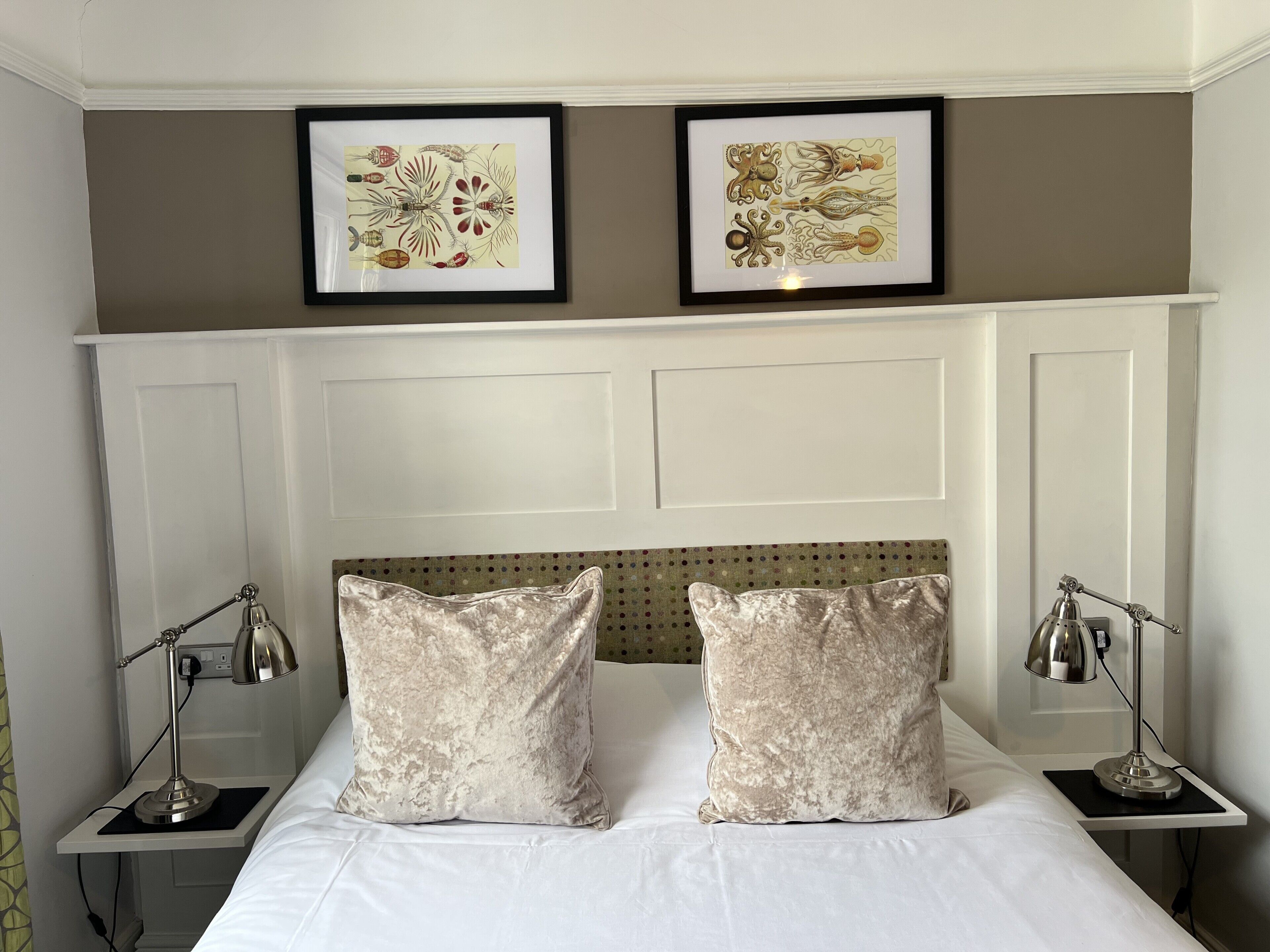 Una cama bien hecha con dos almohadas, cabecera con panel decorativo, dos lámparas de noche y dos obras de arte enmarcadas en la pared.