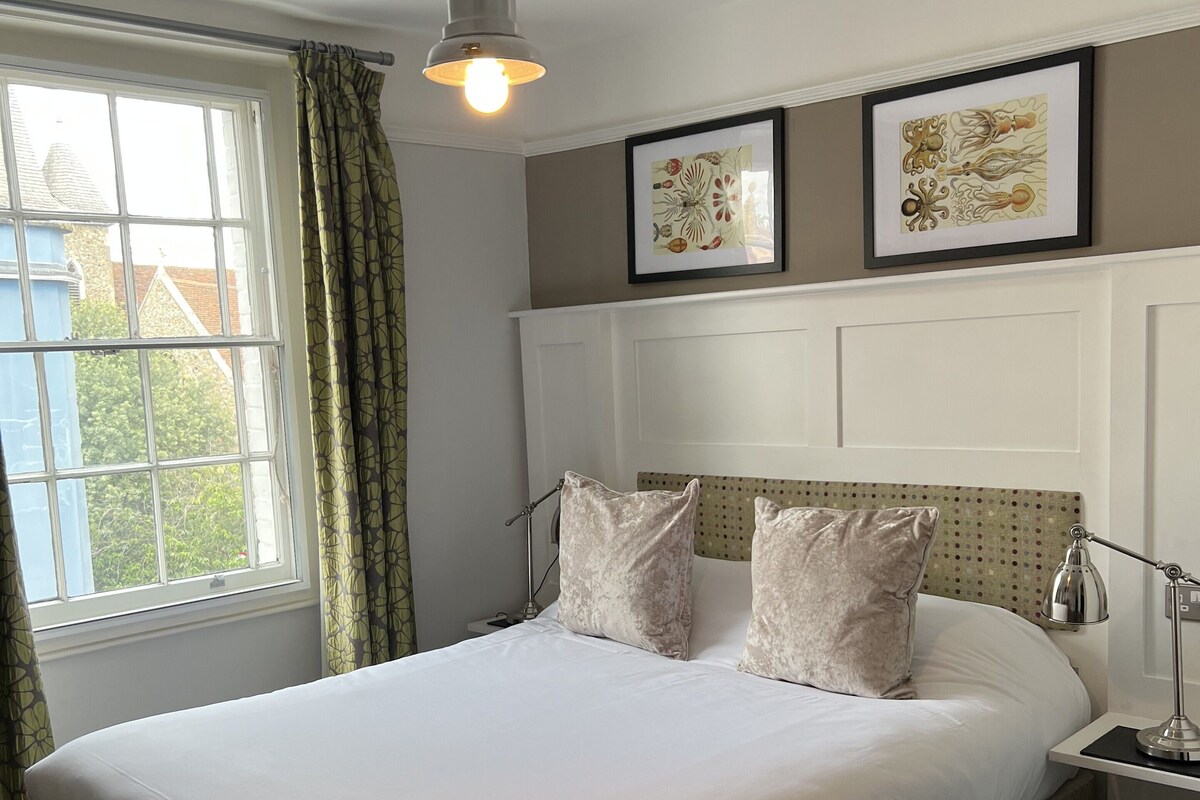 A bedroom with a bed, a window with curtains, a bedside lamp, and two framed pictures on the wall.