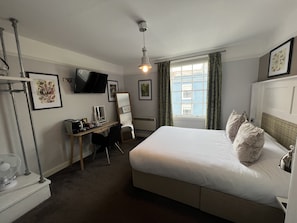 A hotel room with a bed, a desk with a chair, a TV mounted on the wall, and a window with curtains. - The White Horse (Maldon)