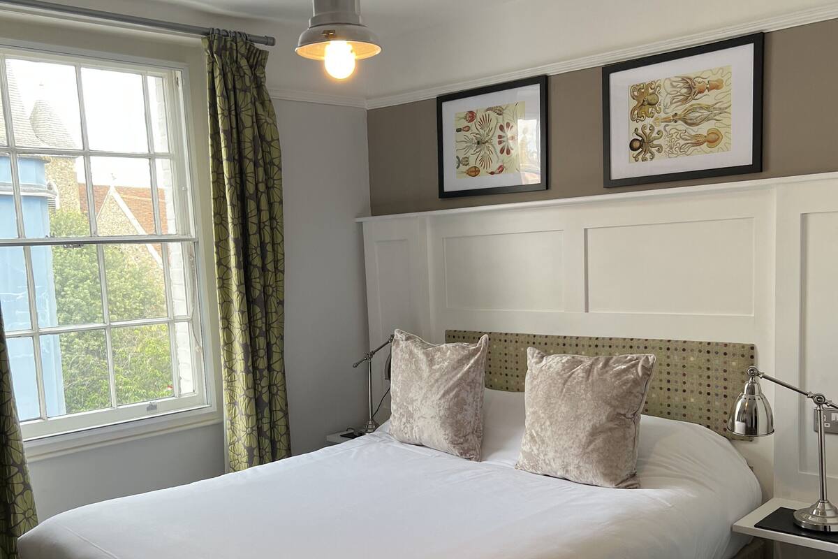 A bedroom with a bed, a window with curtains, a bedside lamp, and two framed pictures on the wall.