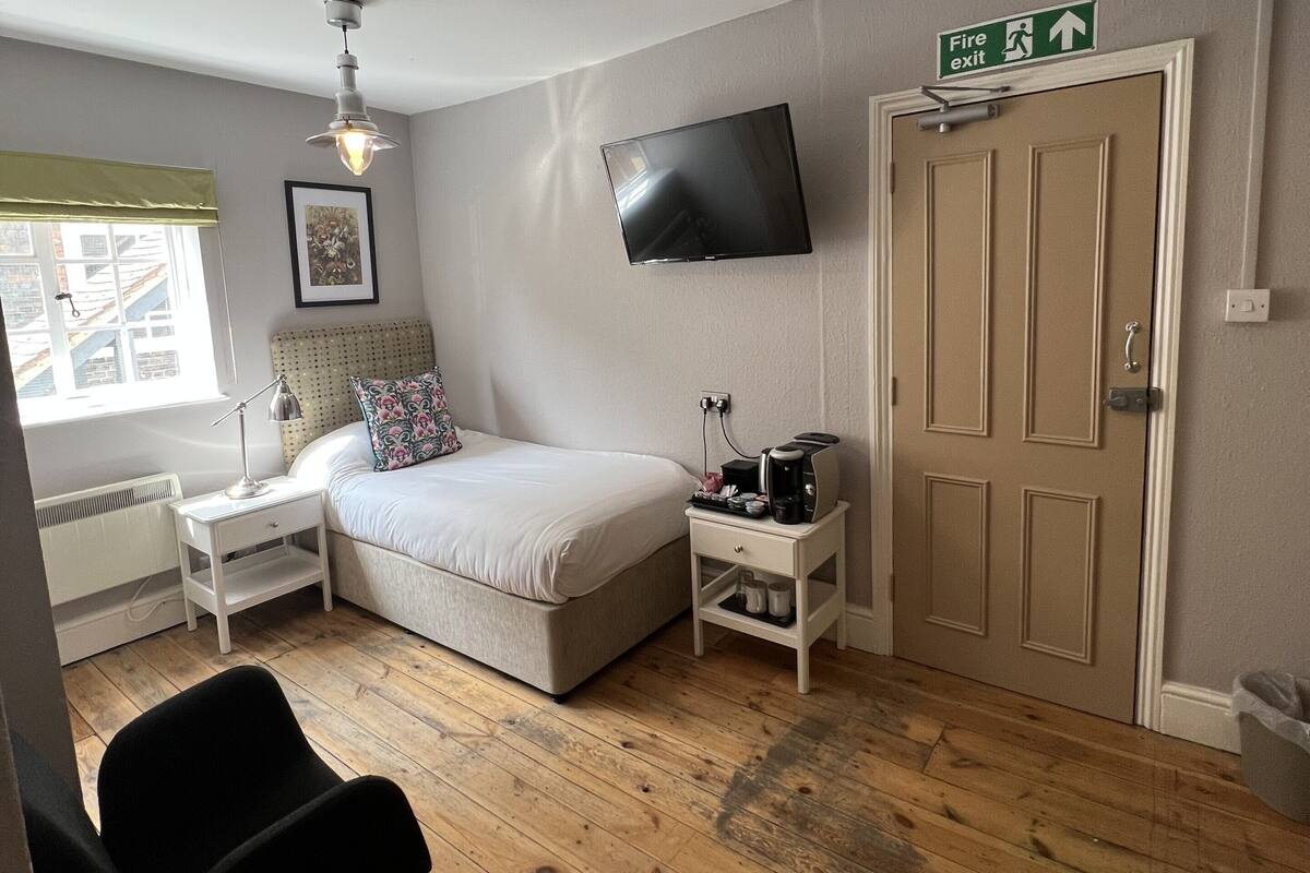 A bedroom with a bed, bedside table, chair, TV, and a door with a fire exit sign.