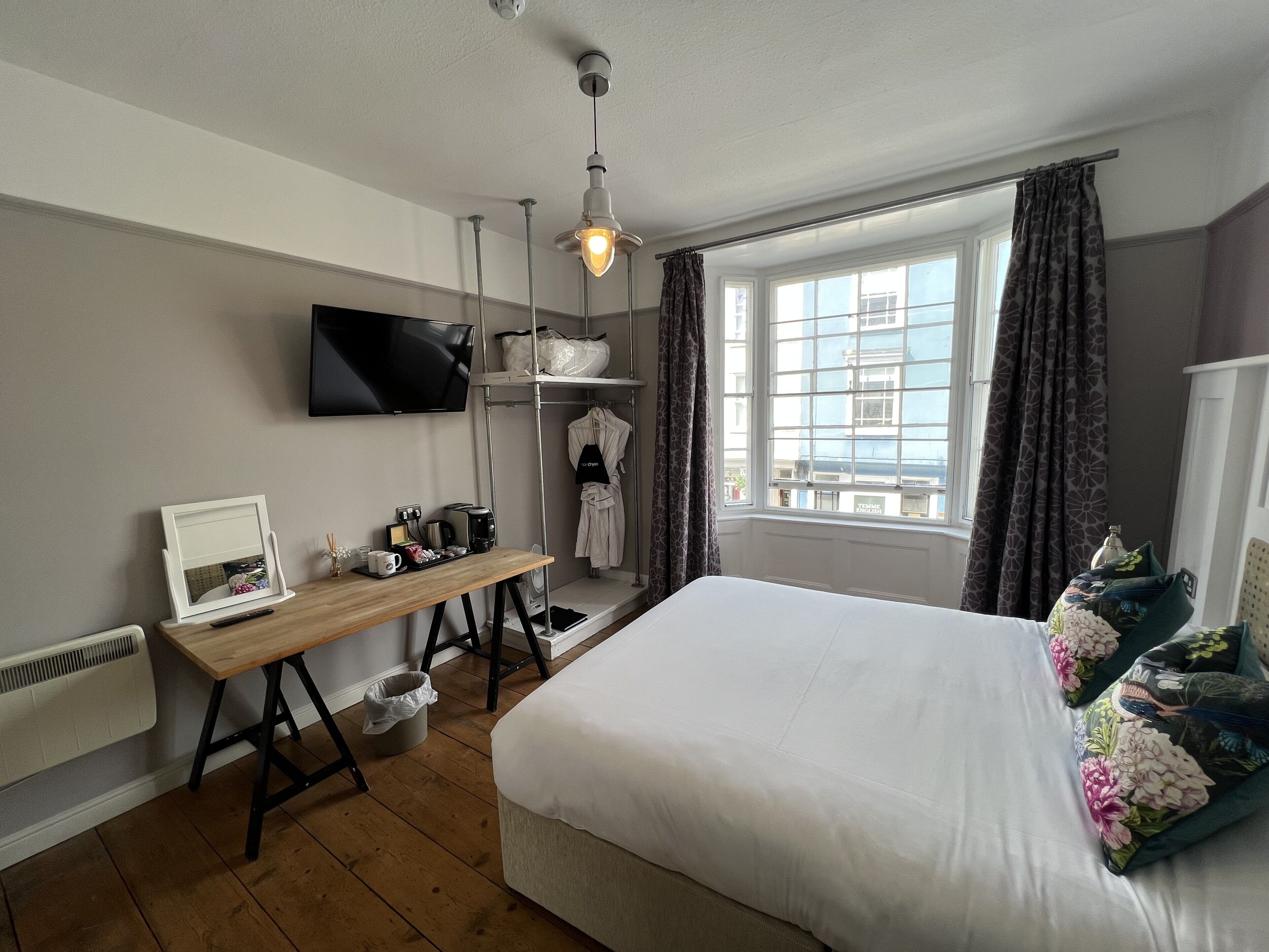 A hotel room with a bed, a desk with a computer, a TV, a window with curtains, and a radiator.
