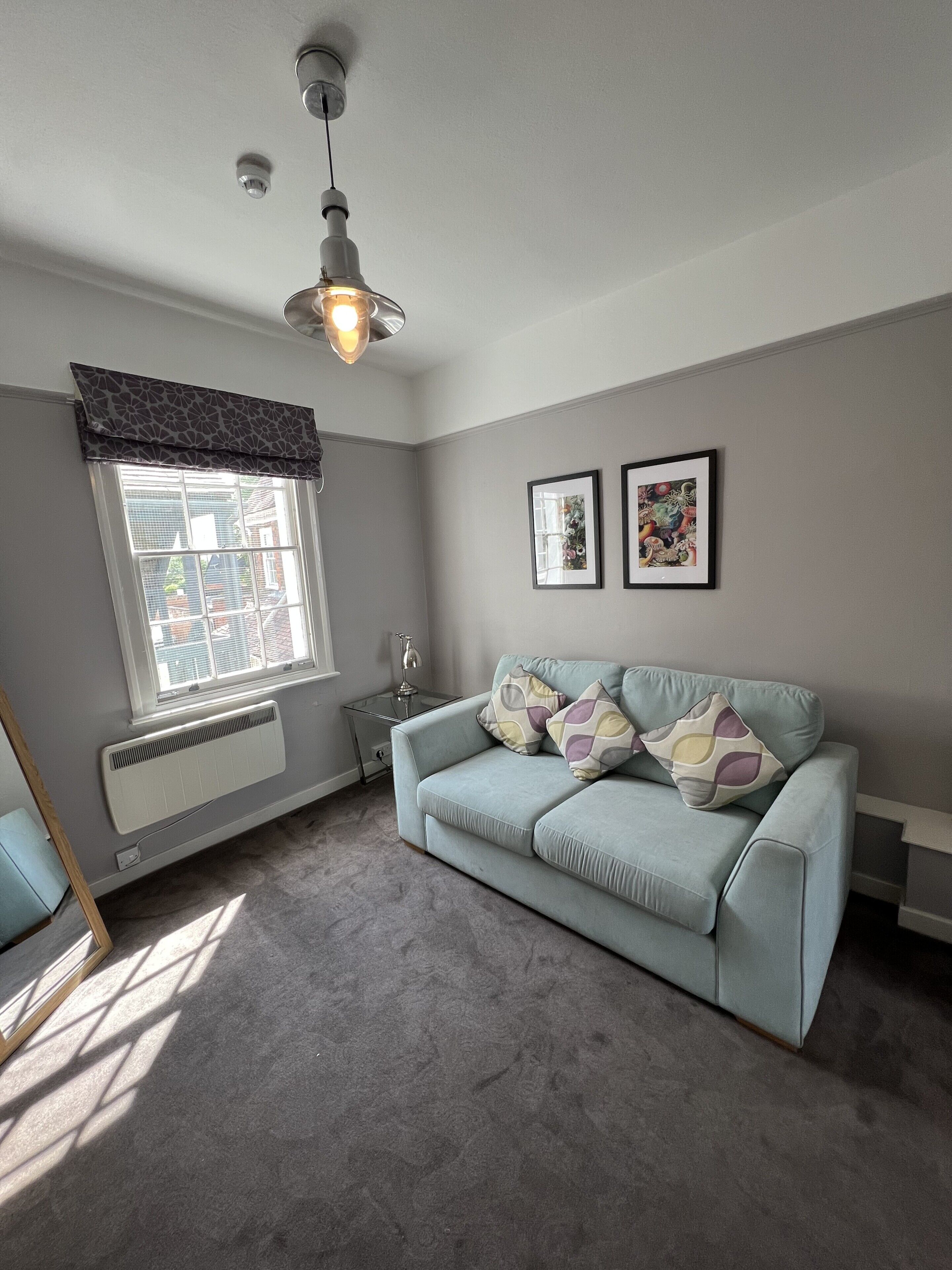 Ein Zimmer mit einem hellblauen Sofa, einem Heizkörper, einem Fenster mit Vorhang, einer Deckenleuchte und zwei gerahmten Bildern an der Wand.