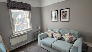 A room with a light blue sofa, a radiator, a window with a curtain, a ceiling light, and two framed pictures on the wall. - The White Horse (Maldon)