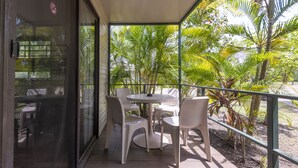Un área de estar al aire libre con techo, con una mesa y sillas, rodeada de exuberante vegetación y palmeras.