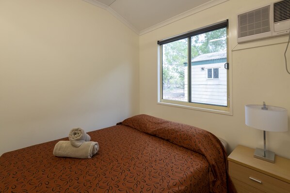 A bedroom with a bed, a window, a bedside table with a lamp, and a wall-mounted air conditioner.