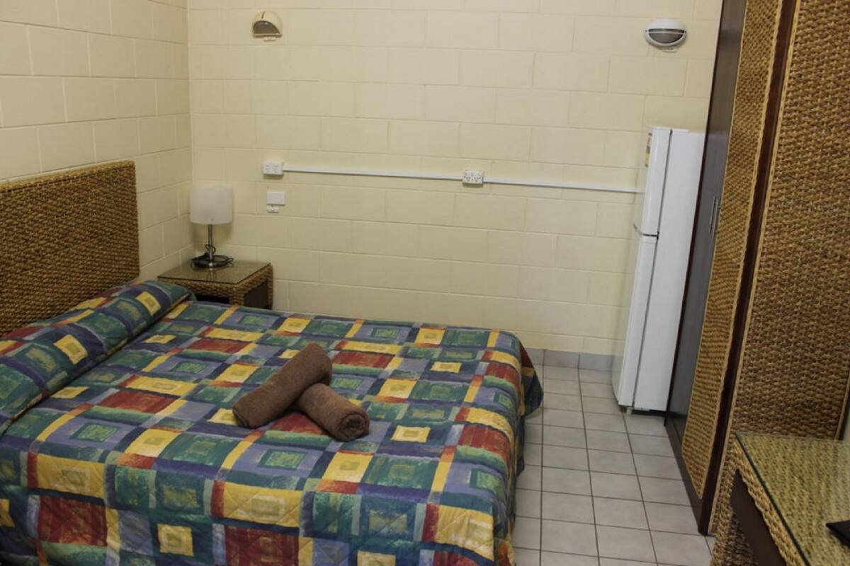 Una habitación pequeña y acogedora con cama, mesita de noche, lámpara y refrigerador.