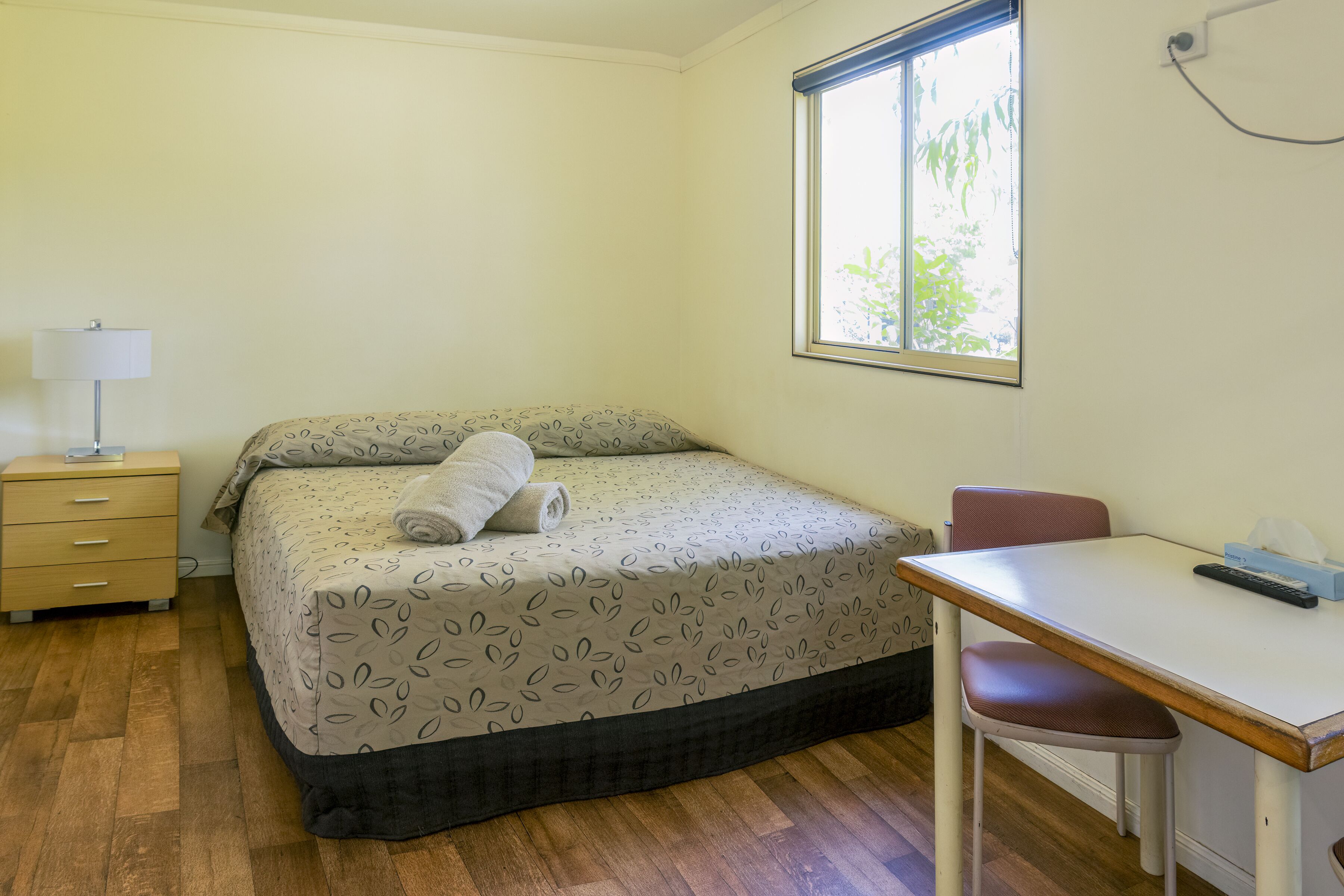 A bedroom with a bed, a desk, a chair, a nightstand, and a window.